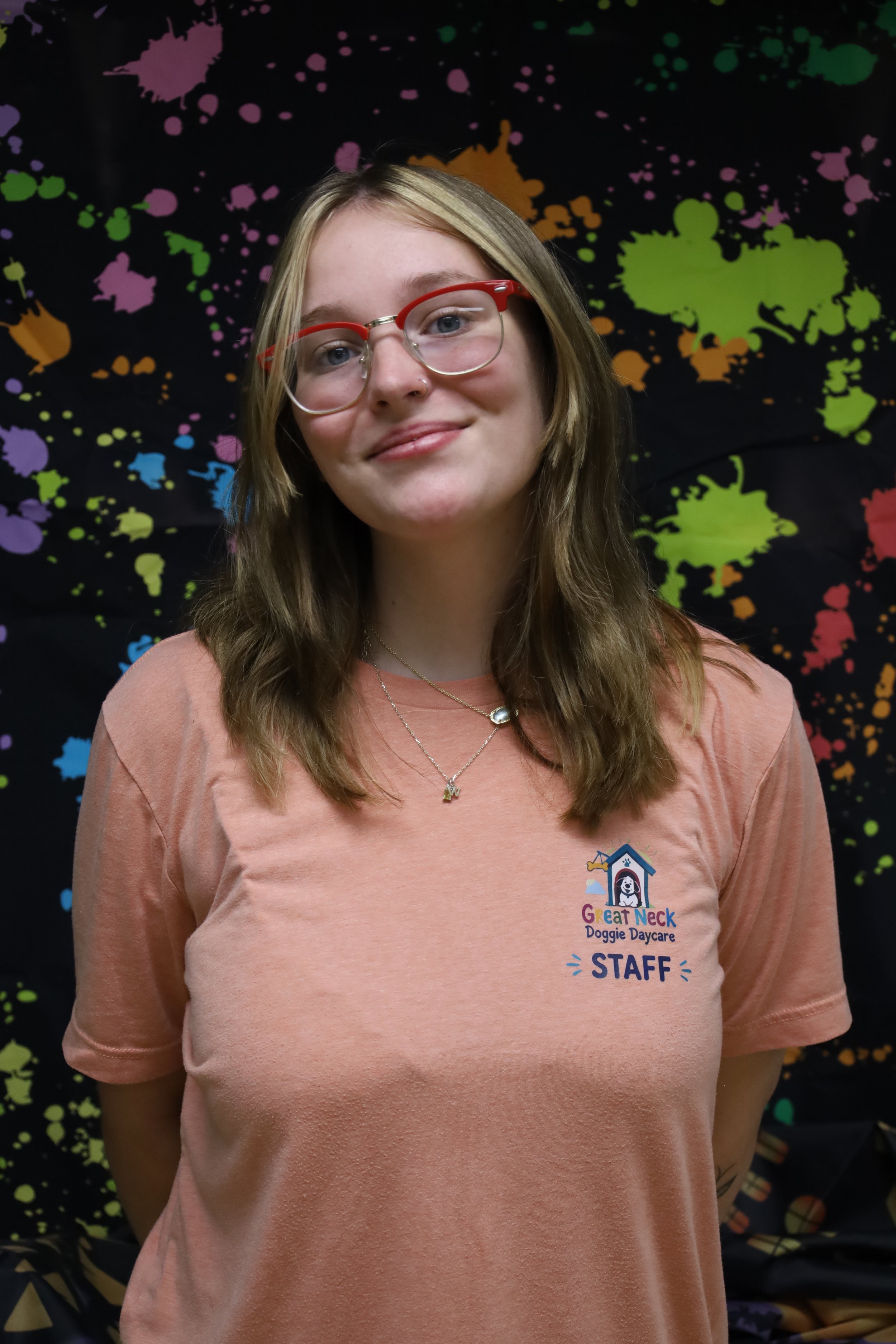 A smiling young woman with glasses and long hair, wearing a peach-colored T-shirt with a staff badge for Great Neck Doggie Daycare, standing in front of a black background with colorful paint splatters.