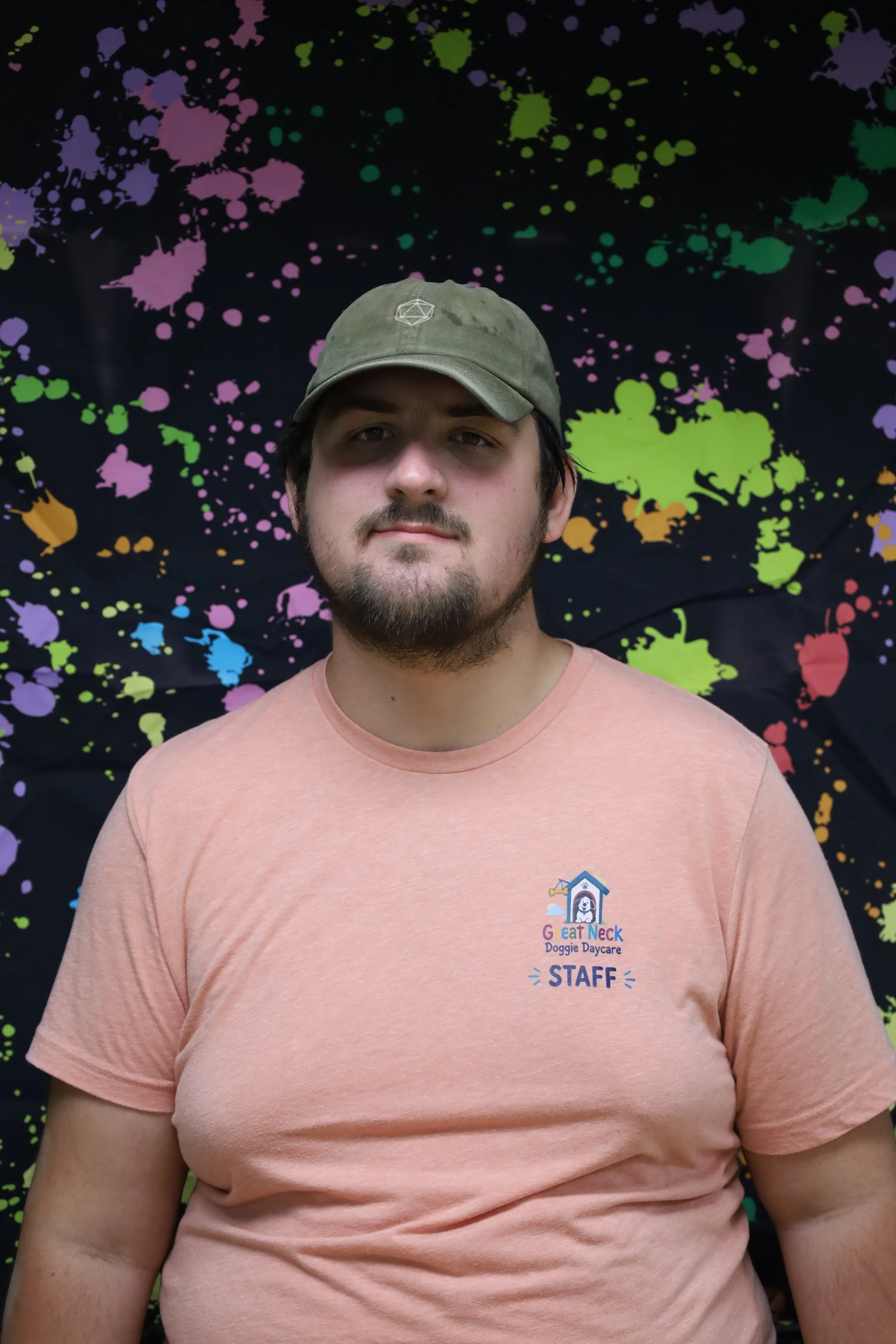 A young man with a light beard and mustache wearing a green cap and a peach T-shirt that says 'Great Neck Doggie Daycare STAFF.' He is standing in front of a black background with colorful paint splatters.