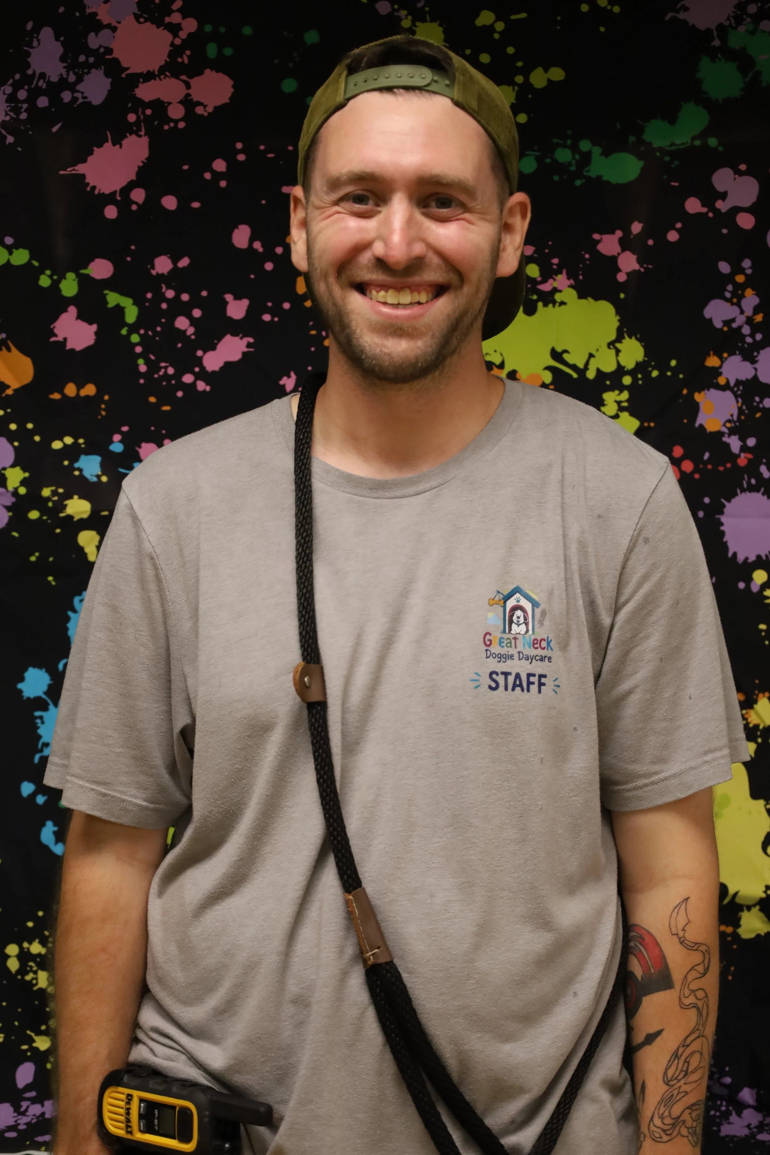 A smiling man wearing a backwards green cap, a gray Great Neck Doggie Daycare staff t-shirt, and a lanyard, standing in front of a colorful, splattered paint background.