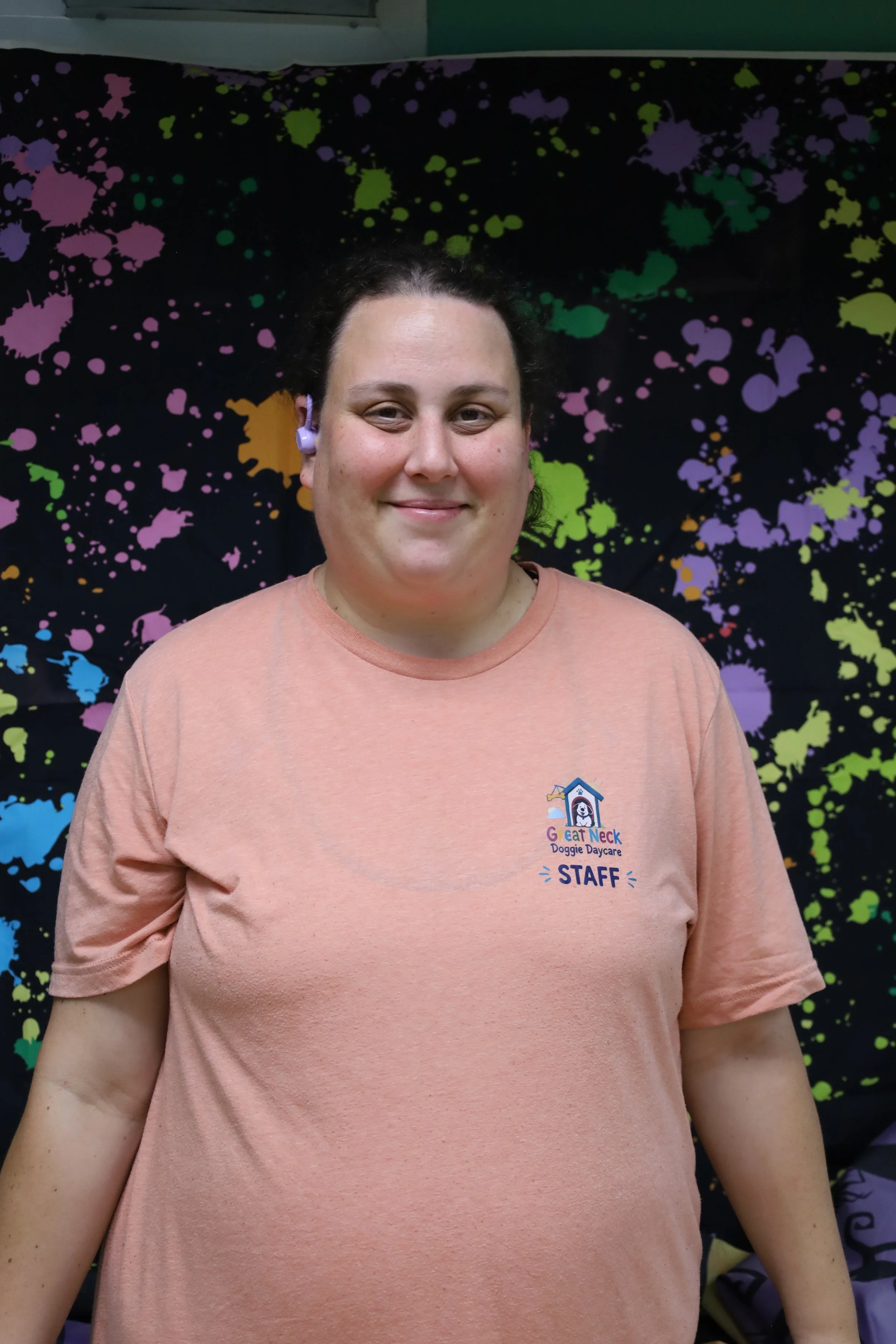 A woman smiling, wearing a peach-colored T-shirt with a logo that reads 'Great Neck Doggie Daycare STAFF', standing in front of a colorful splatter paint backdrop.
