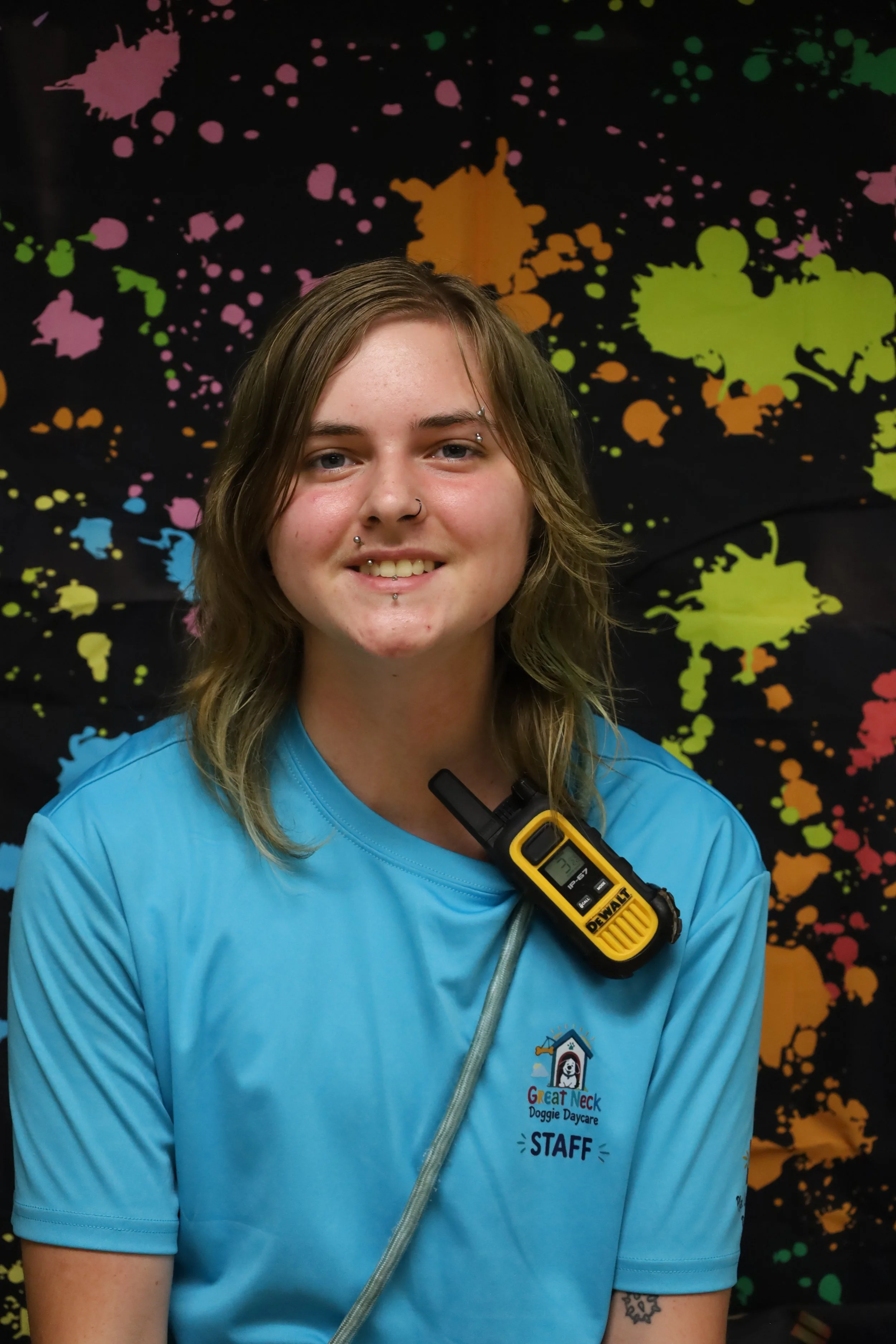 Happy young woman wearing a bright blue shirt with a logo that reads 'Great Neck Doggie Daycare' and a staff badge. She has shoulder-length light brown hair, a nose piercing, and a lip piercing, and is standing in front of a colorful, splattered paint background.