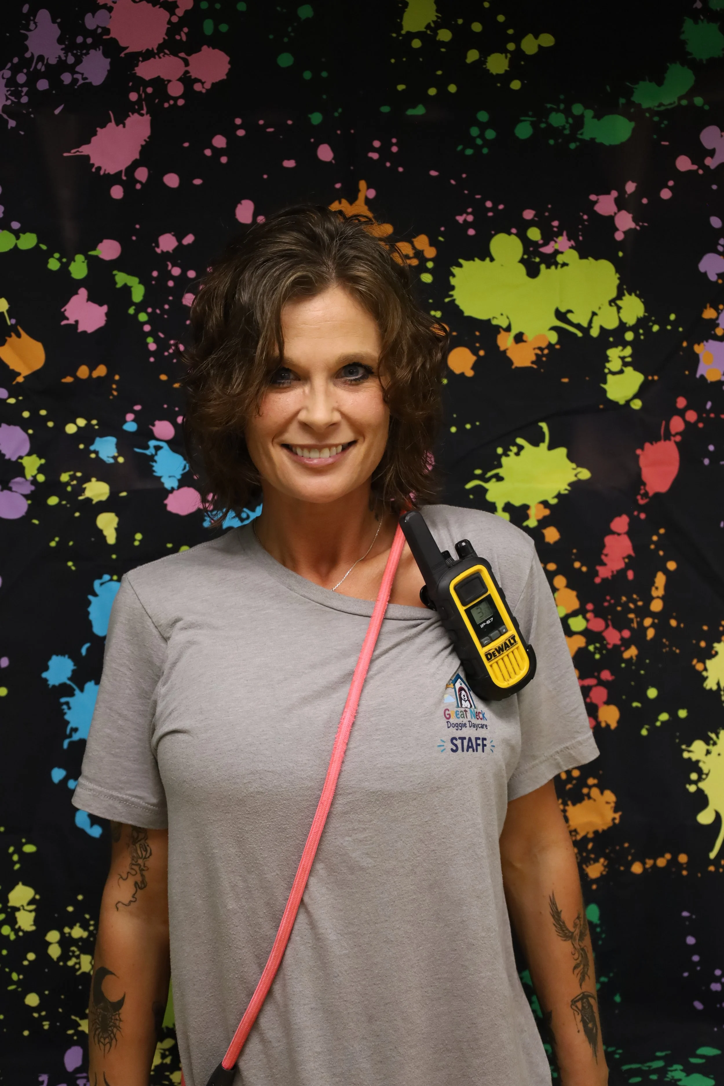 A smiling woman with short wavy brown hair, wearing a gray T-shirt with a logo and the word 'STAFF', standing in front of a colorful splatter paint background. She has tattoos on her arms and is holding a walkie-talkie.