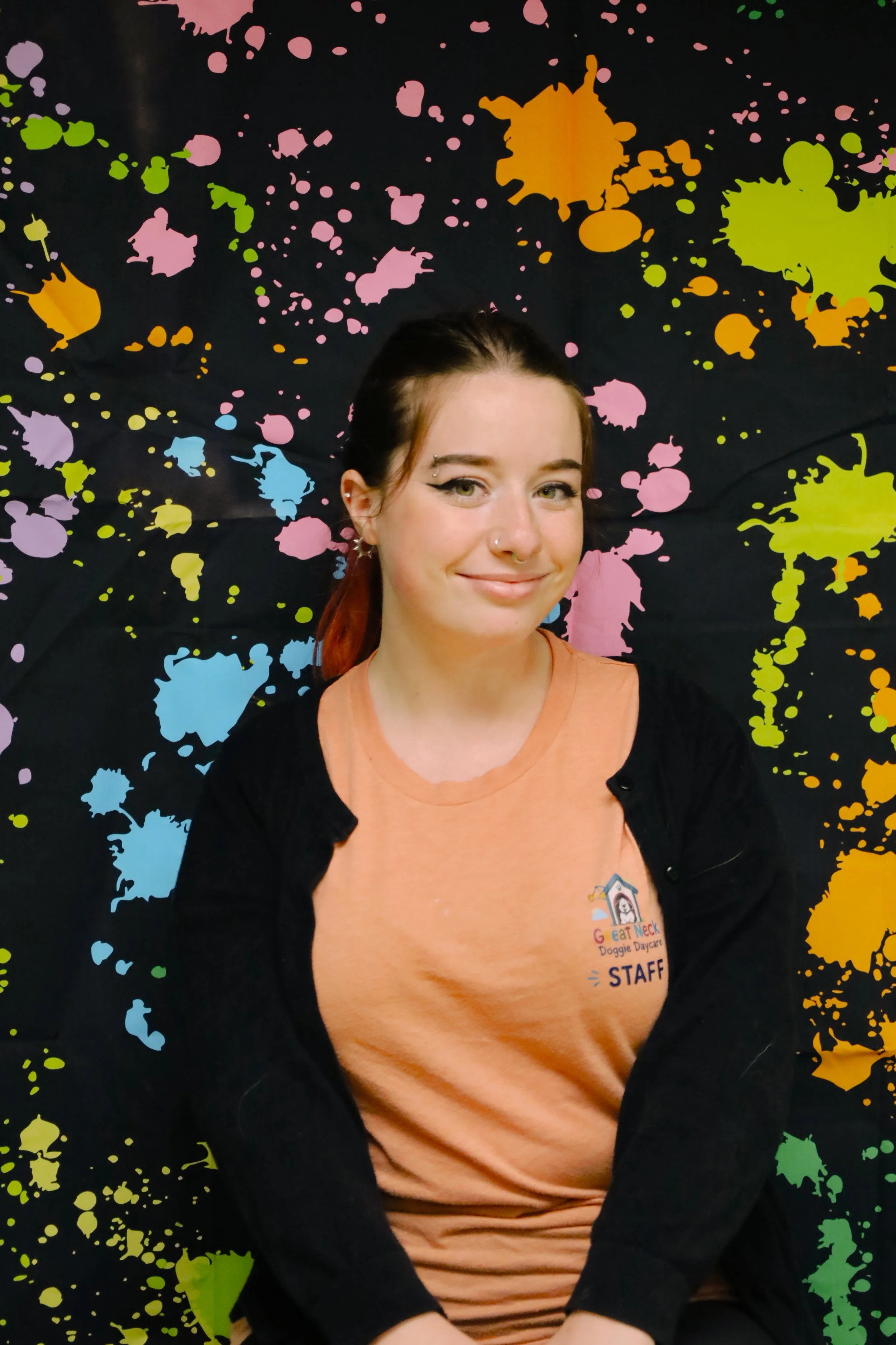 A young woman with light skin, dark hair tied back, and wearing makeup, smiles at the camera. She is wearing a peach-colored shirt with a logo and text that reads "G Eat Neak Doggie Daycare STAFF." She is sitting in front of a black background with colorful splatter paint in pink, orange, yellow, green, blue, and purple.