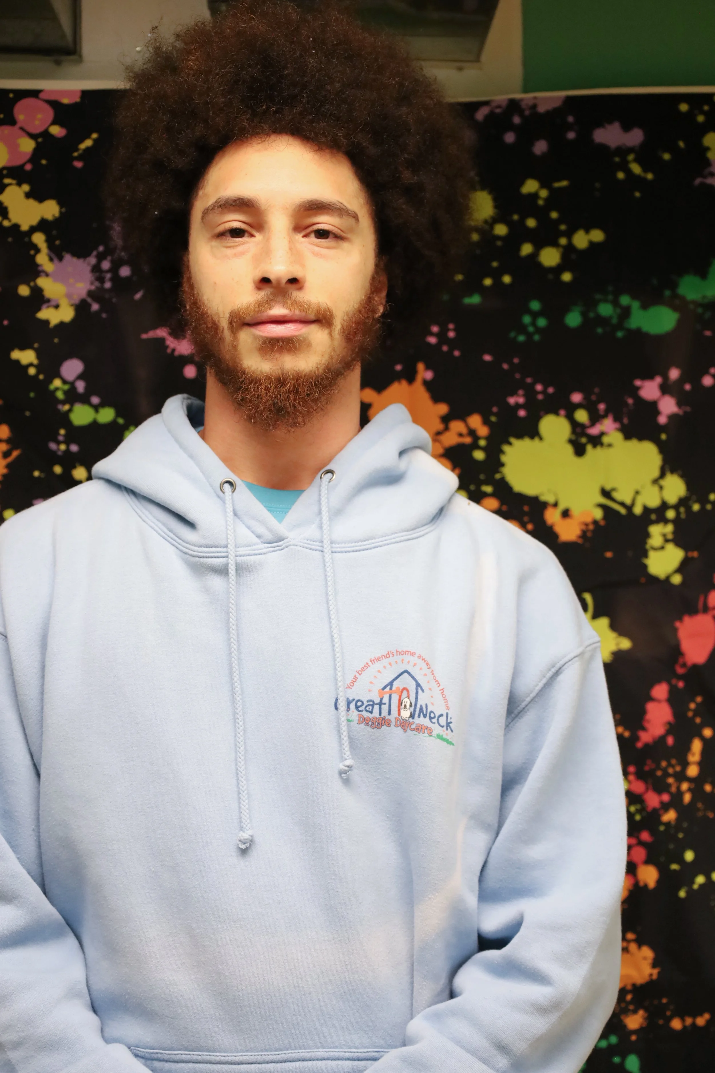 A young man with curly hair and a beard wearing a light blue hoodie with a logo on it, standing in front of a colorful splatter paint backdrop.