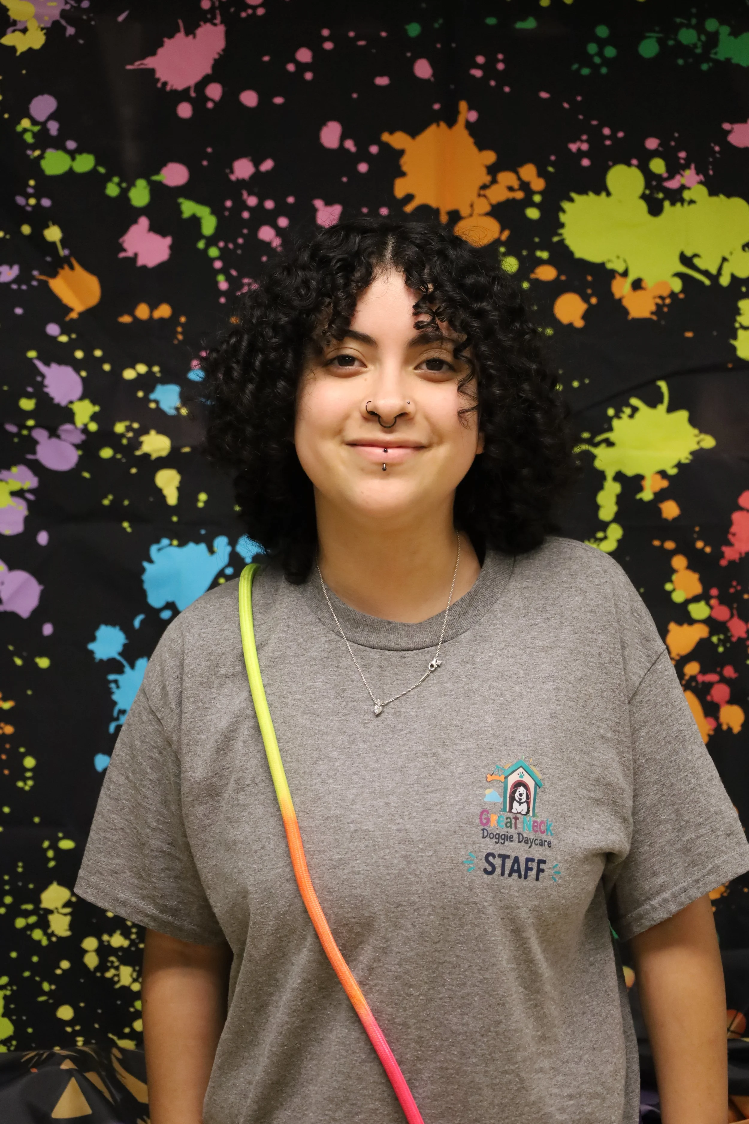 A woman with curly black hair and nose piercings smiling in front of a black background with colorful paint splashes, wearing a gray staff shirt at Doggie Daycare.