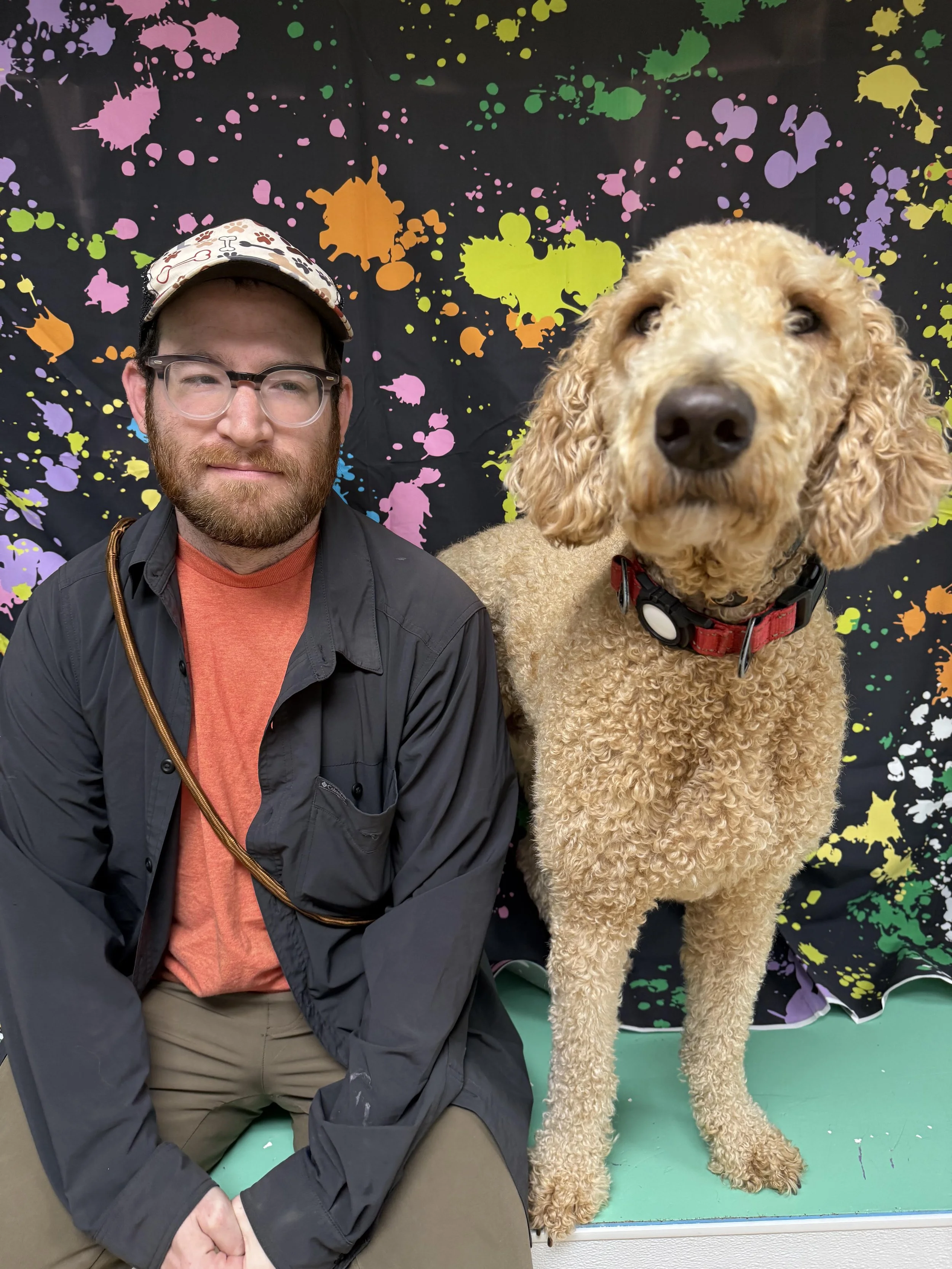 A man with glasses, a beard, and wearing a cap with a pattern, sitting beside a large, curly-haired dog with a red collar, in front of a colorful, splatter-painted backdrop.