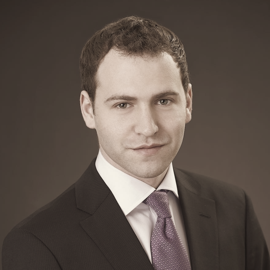 A young man with short brown hair in a dark suit and light-colored shirt, looking at the camera against a plain dark background.