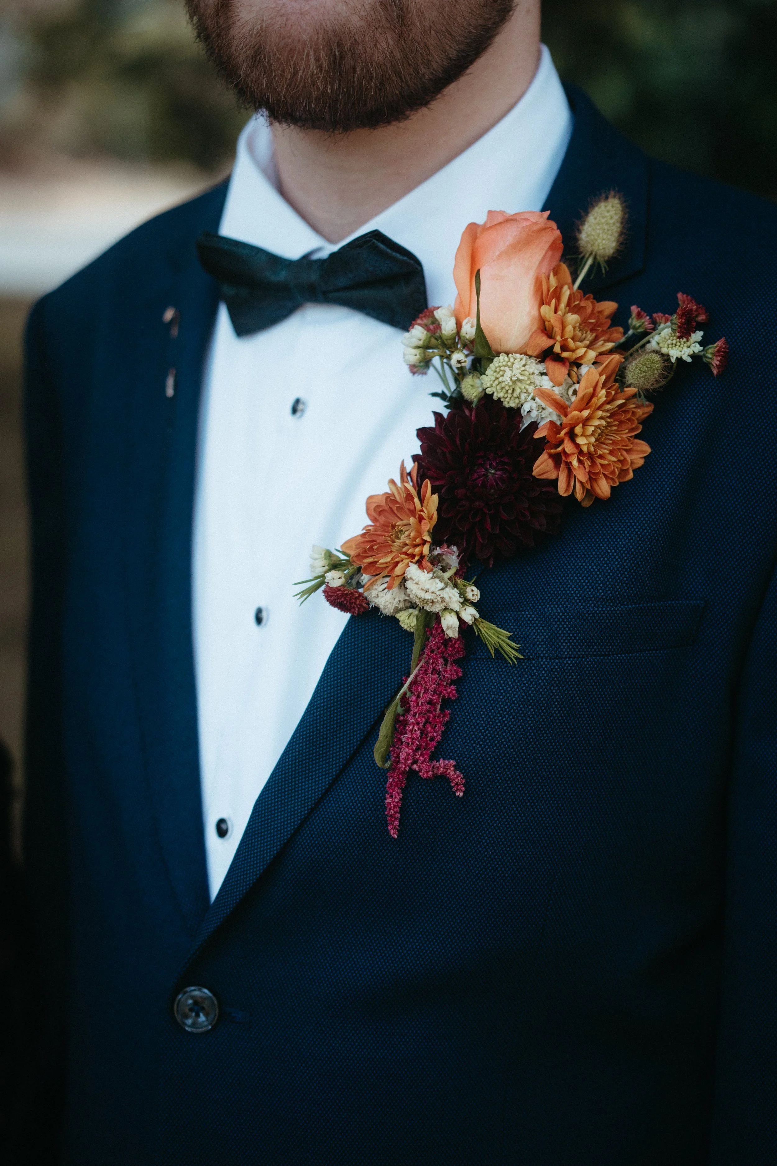 Close-up of a man wearing a formal dark navy tuxedo with a bow tie, adorned with an orange and red floral boutonniere on his left lapel.