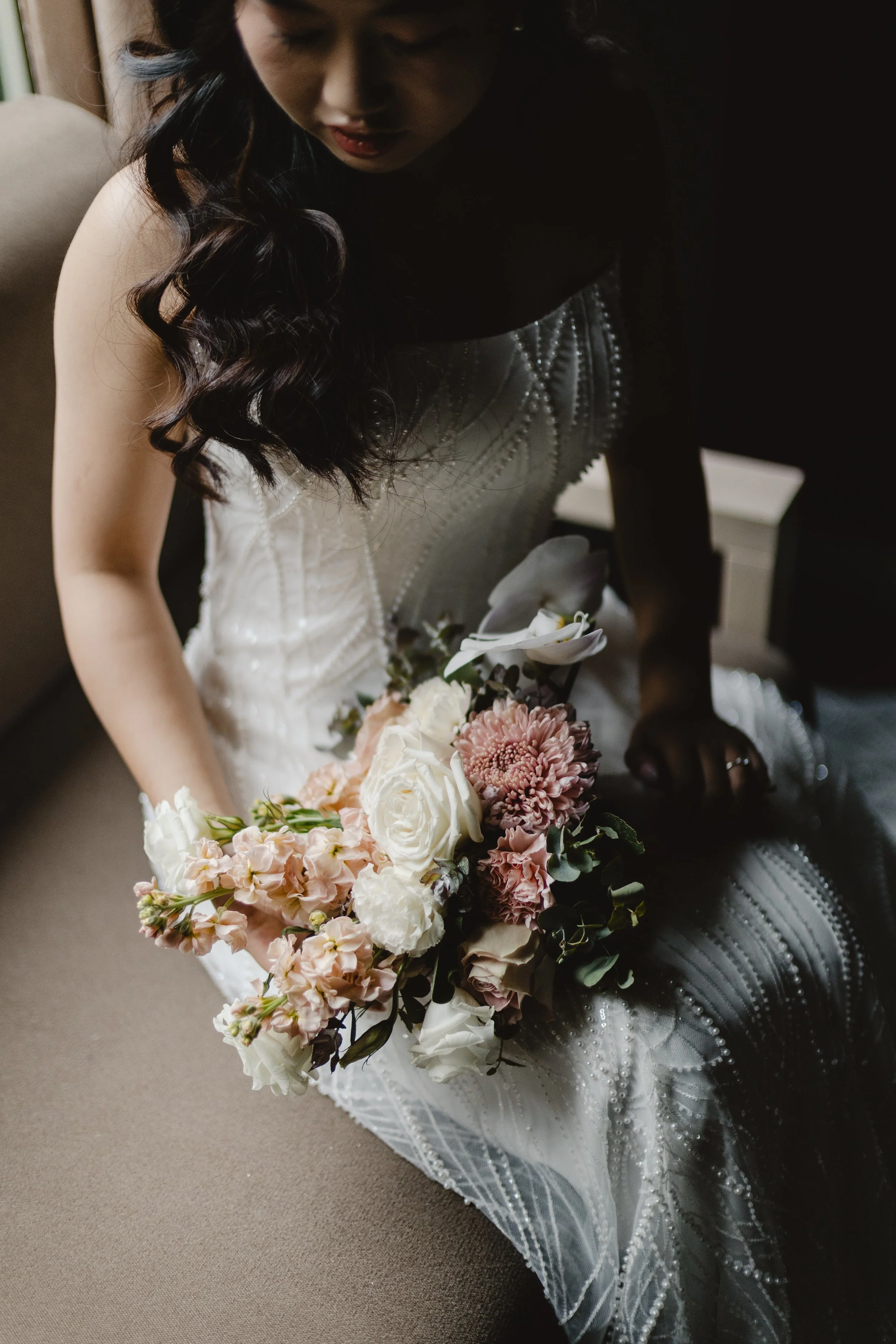 A woman wearing a white wedding dress with pearl and bead embellishments, holding a bouquet of pink, white, and cream flowers. She is bending down or looking at the flowers.