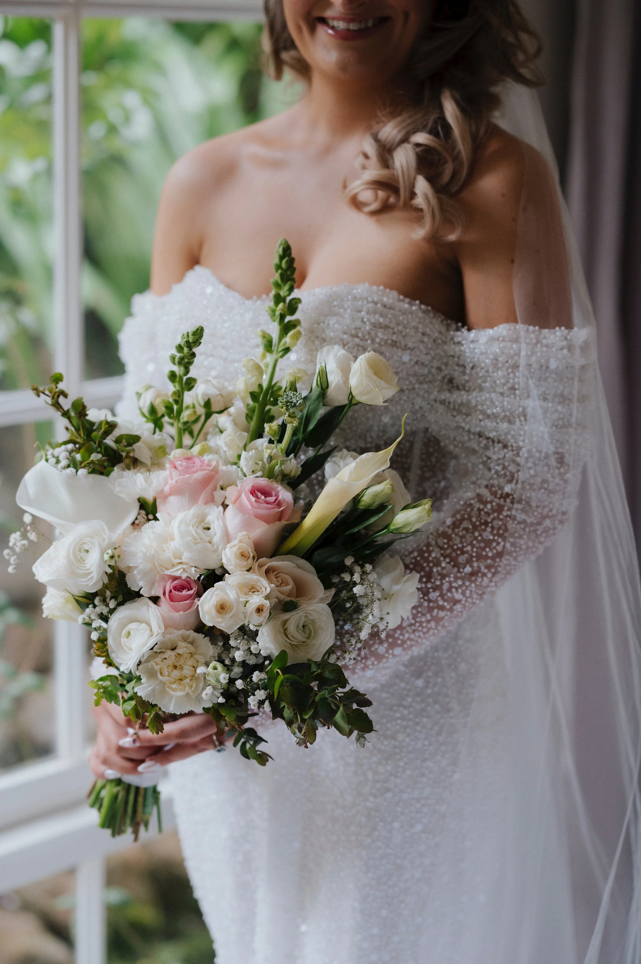 A woman in a wedding dress holding a bouquet of white and pink flowers, standing indoors in front of a window with greenery outside, smiling.