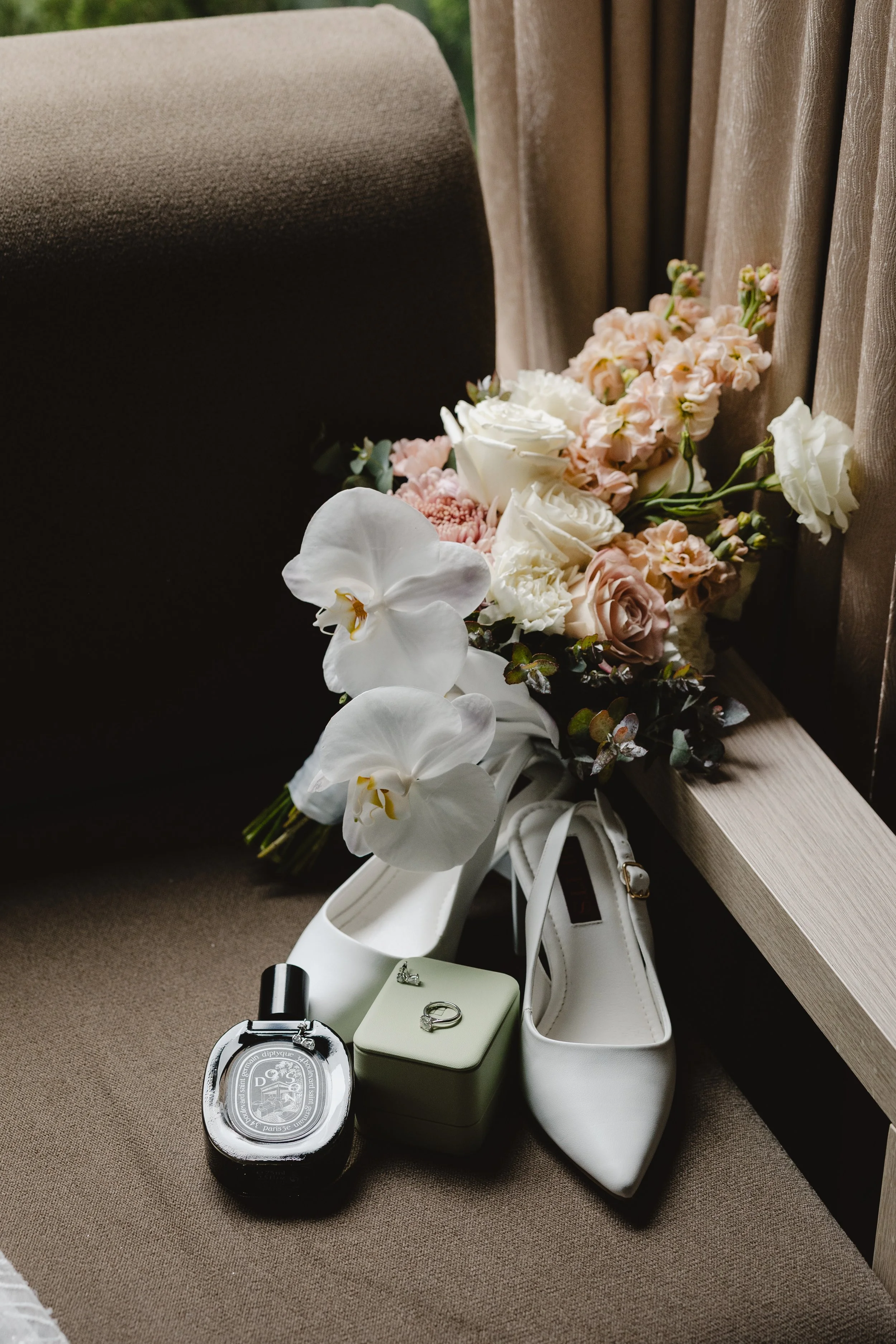 A bouquet of white, pink, and peach-colored flowers rests on a beige fabric surface, with a pair of white high-heeled shoes, a jewelry box with wedding rings, and some small gift boxes nearby.