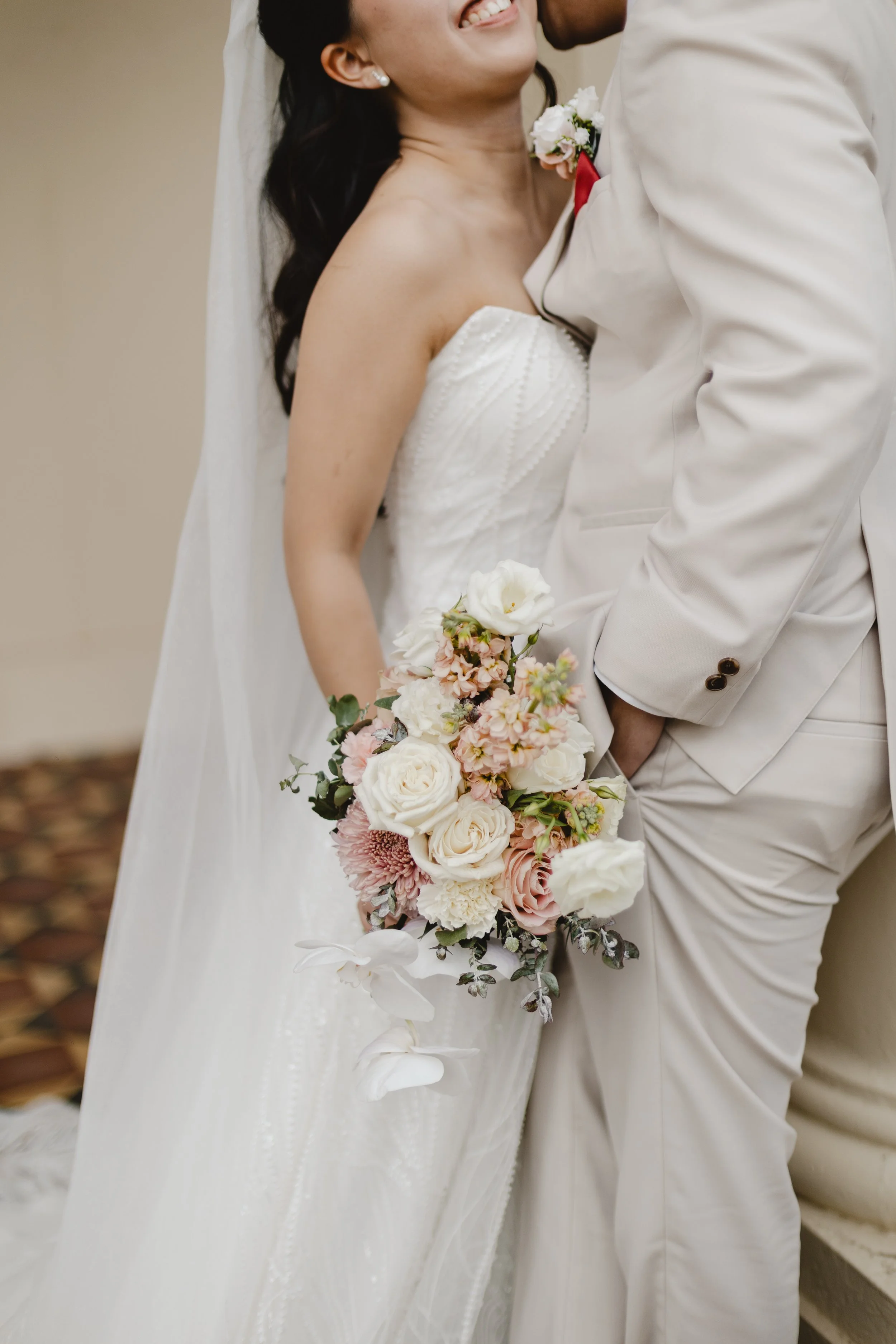 A bride and groom share a moment, with the bride holding a bouquet of white and pink flowers. The bride is smiling, wearing a white strapless wedding dress, and has dark hair. The groom is dressed in a light-colored suit with a white shirt and a red boutonniere.