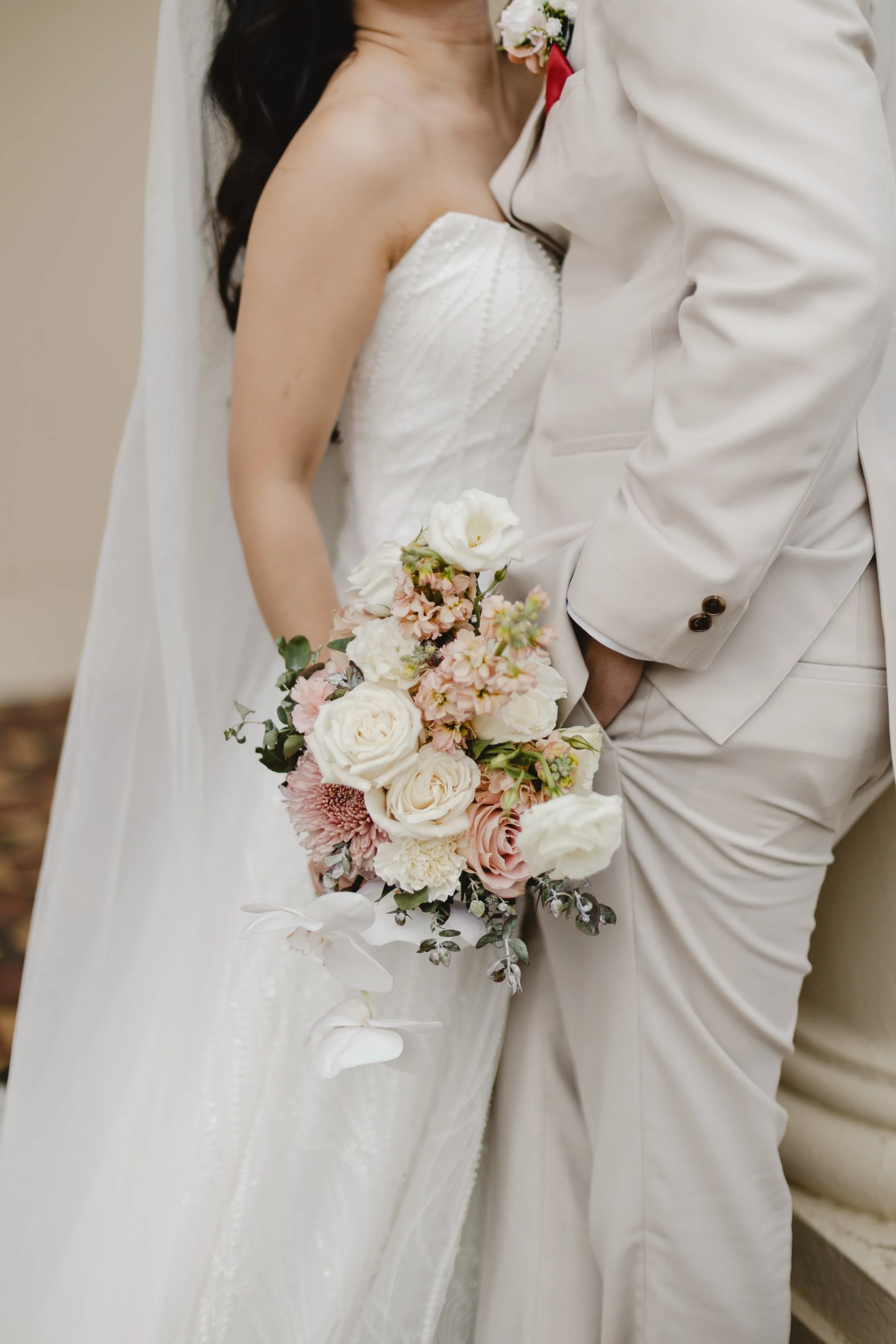 Close-up of a bride and groom holding a bouquet of white, pink, and blush flowers on their wedding day. The bride is wearing a strapless white wedding dress, and the groom is in a light beige suit.