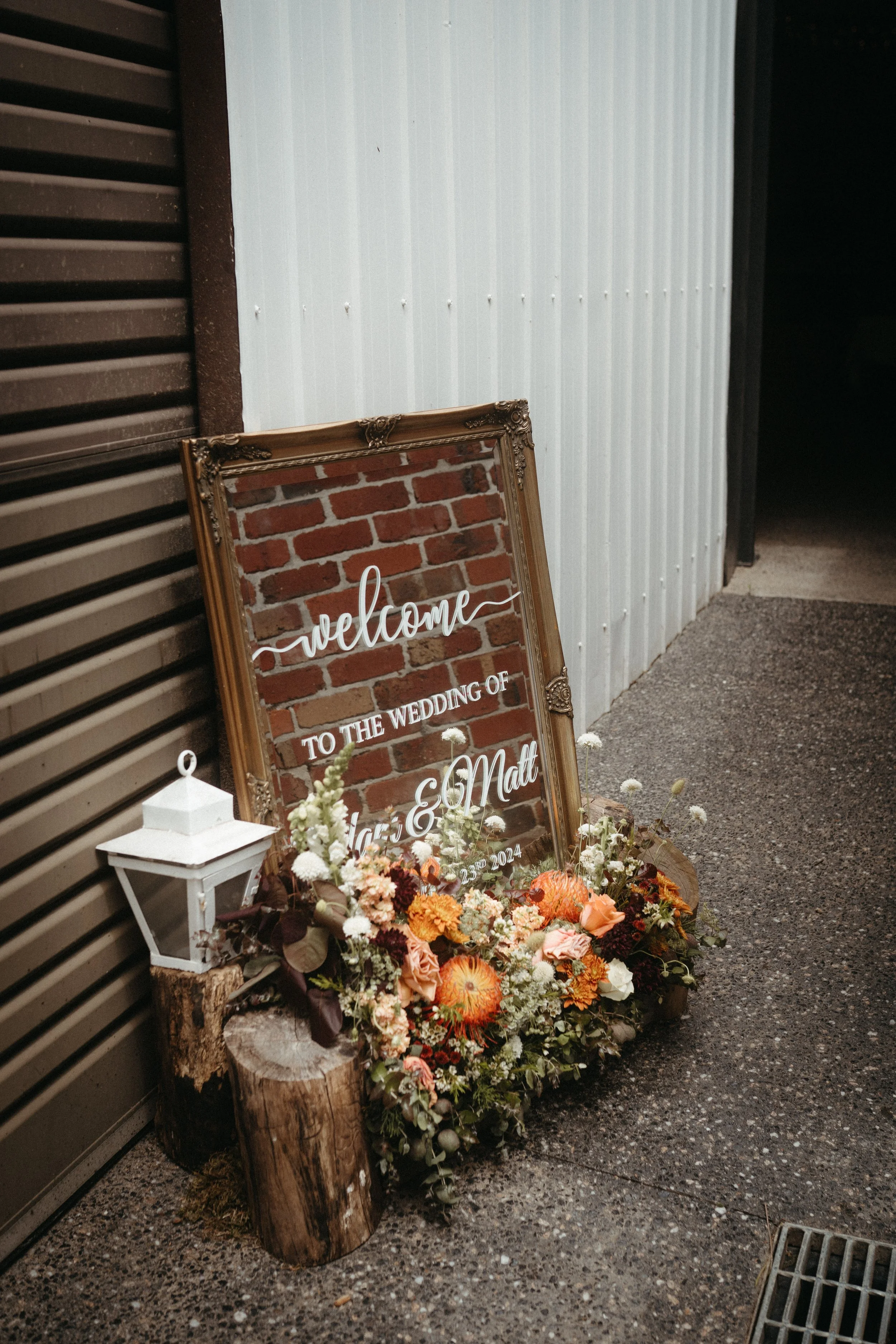 Decorative wedding sign with floral arrangement, lantern, and wooden log in front of a brick and metal wall, welcoming guests to the wedding of a couple.