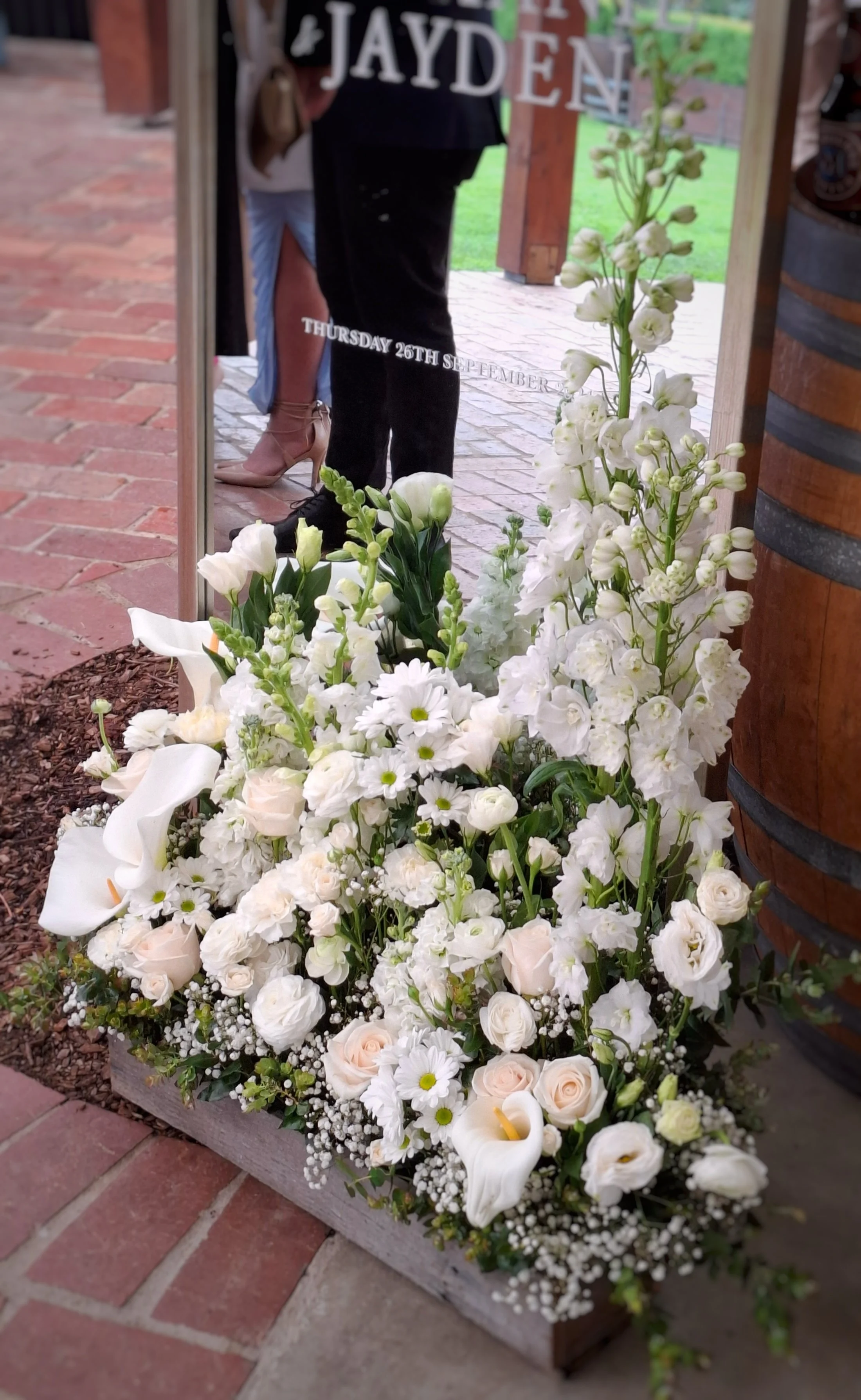 A large arrangement of white flowers, including roses and calla lilies, in a wooden container outside near a brick pathway and a wooden barrel.