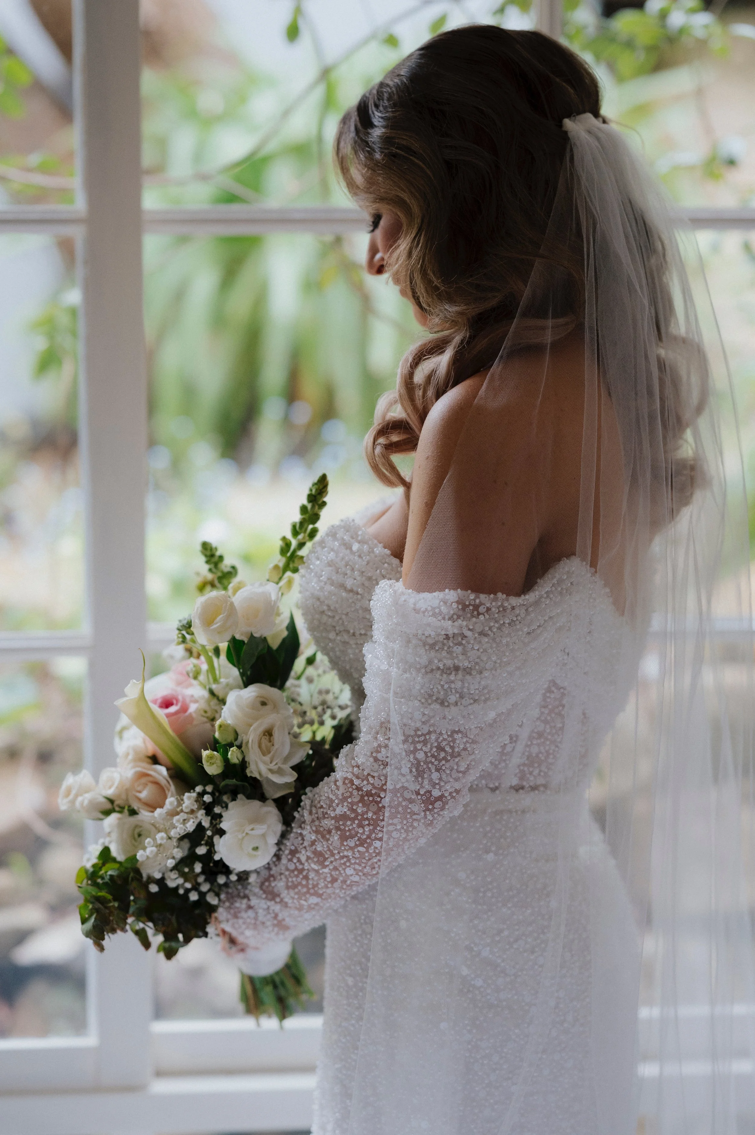 A bride in a white wedding dress holding a bouquet of white and blush pink roses, standing by a window with a scenic outdoor view, her face turned downward with a veil in her wavy brown hair.