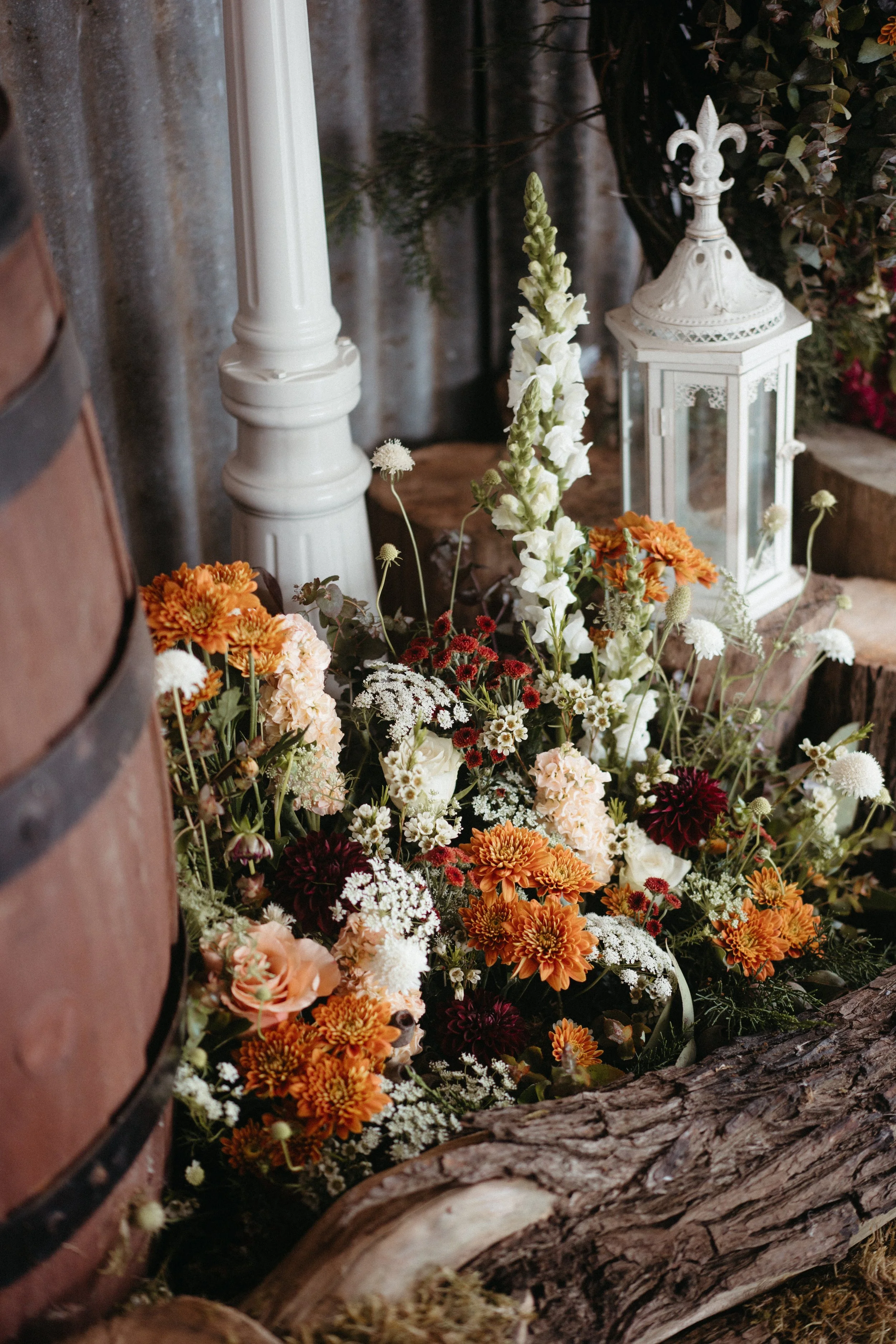 Decorative arrangement of white and orange flowers next to a white lantern and a white candle holder, with logs and greenery in the background.