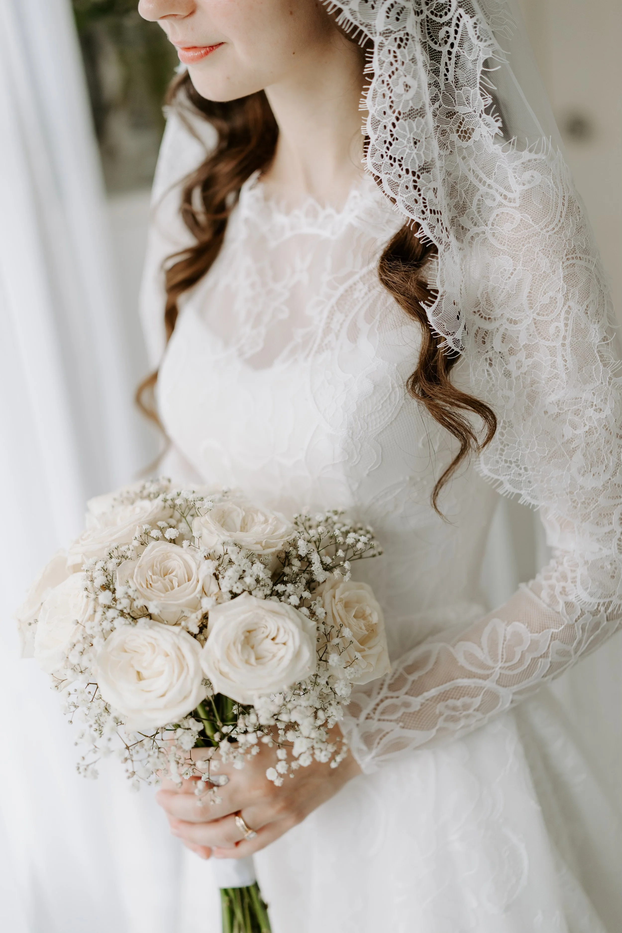 Close-up of a bride holding a bouquet of white roses and baby's breath, wearing a lace wedding dress and veil.