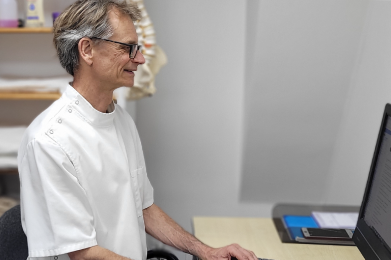 A medical professional, wearing white scrubs, working on a computer in a clinical setting.
