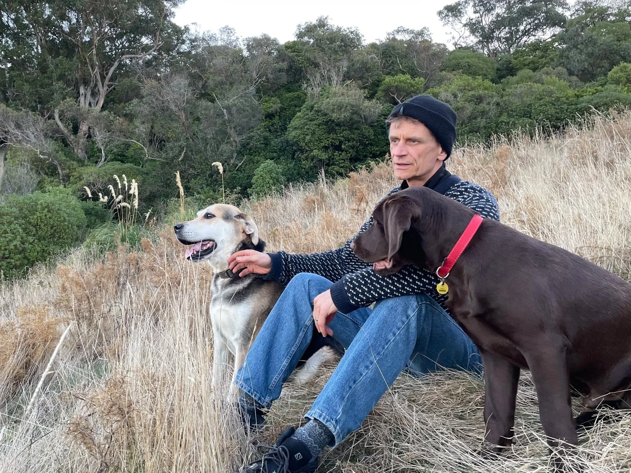 A man sitting outdoors on a grassy hillside holding two dogs, one beige and white, the other dark brown, with a background of trees and bushes.