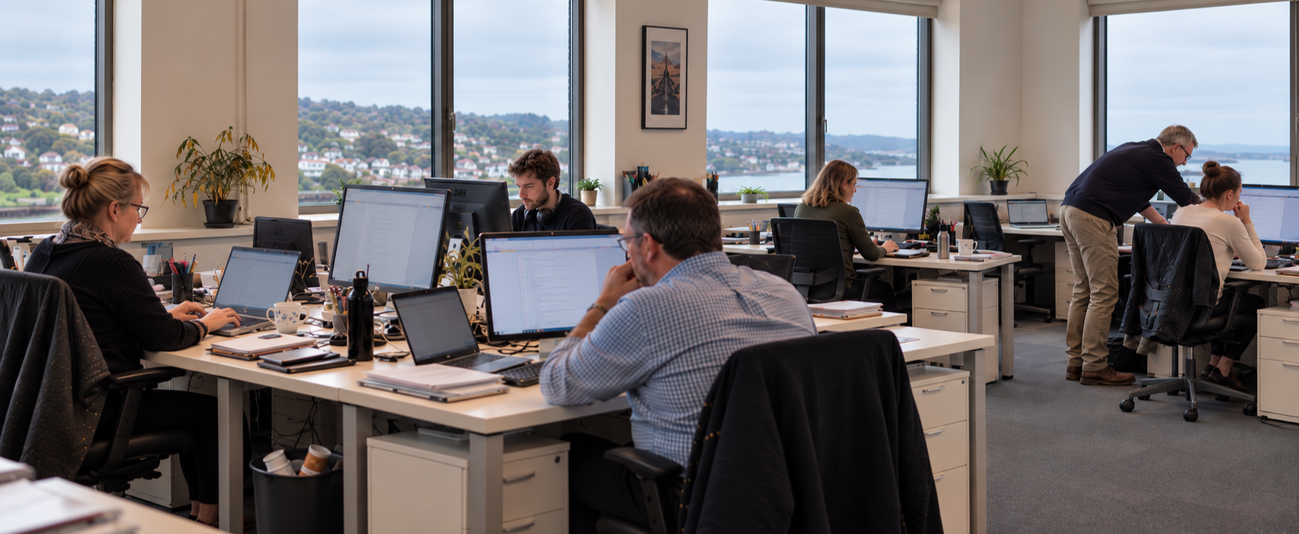Office with multiple employees working at desks with computers, large windows showing a hilly landscape, and indoor plants.