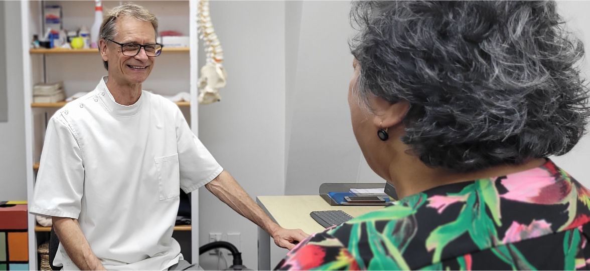 An Osteopath with glasses smiling and sitting in a consultation room, talking to a female patient with curly grey hair wearing a colourful floral shirt.