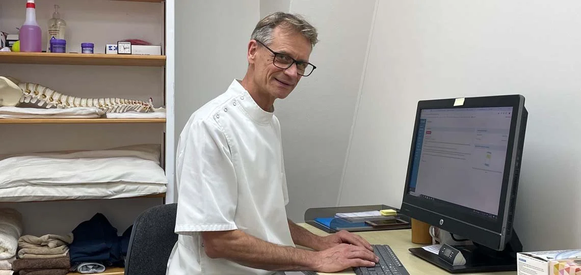 A healthcare professional, wearing a white coat and glasses, working at a desk with a computer in a medical office or clinic setting.