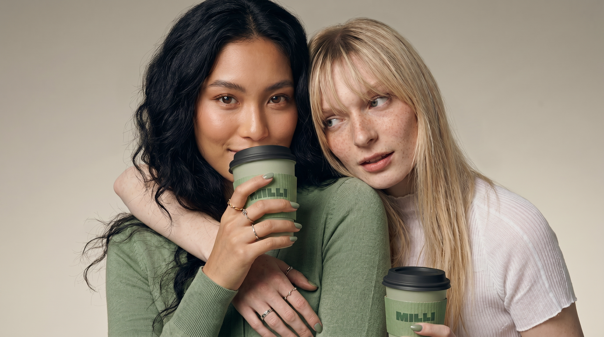 Two young women, one with black hair and the other with blonde hair and freckles, holding coffee cups and close together, smiling softly, against a plain wall background.