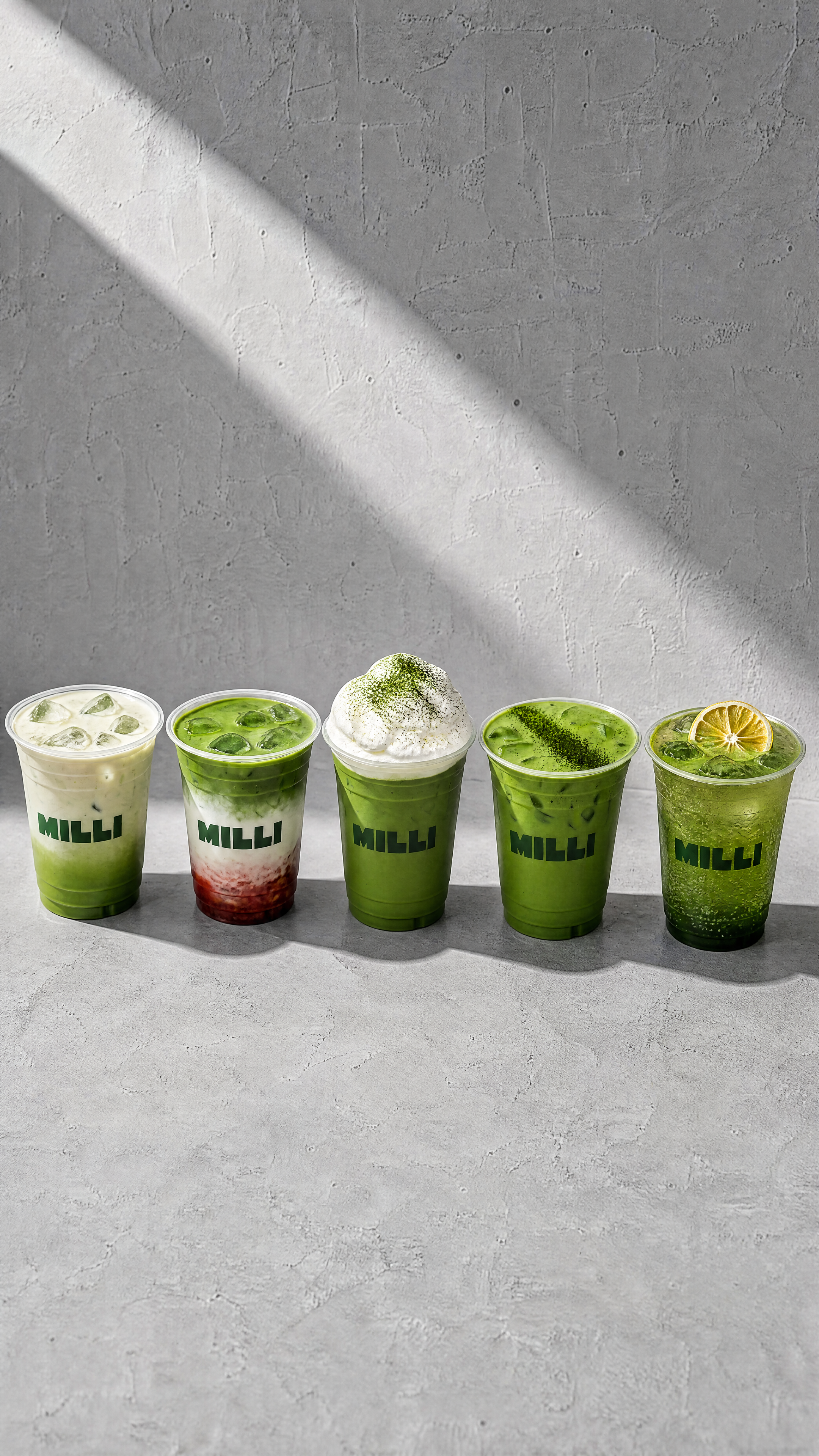 Five iced green beverages in clear cups, each with the word 'MILLI' printed on them, arranged in a row on a gray surface against a gray wall. The drinks vary in appearance, with some containing lemon slices, whipped cream, or layered ingredients.