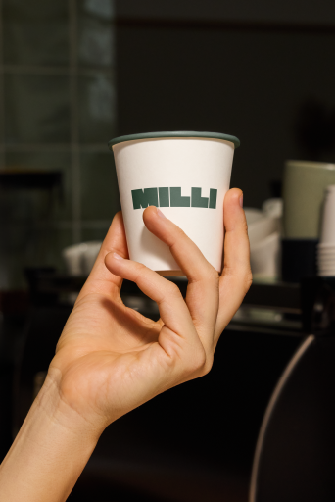 A person holding a white paper cup with green text on it, in an indoor setting with black and green objects in the background.