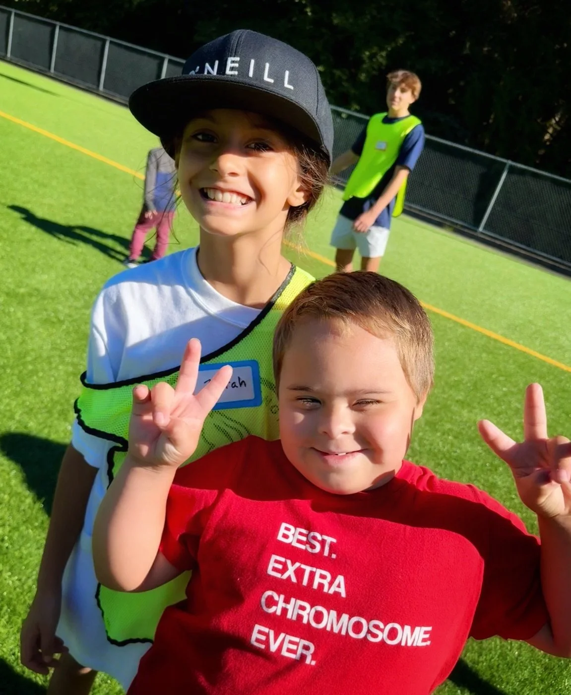 Two smiling kids on a sports field, one in a cap and a bright vest, one in a red shirt, both holding up peace signs