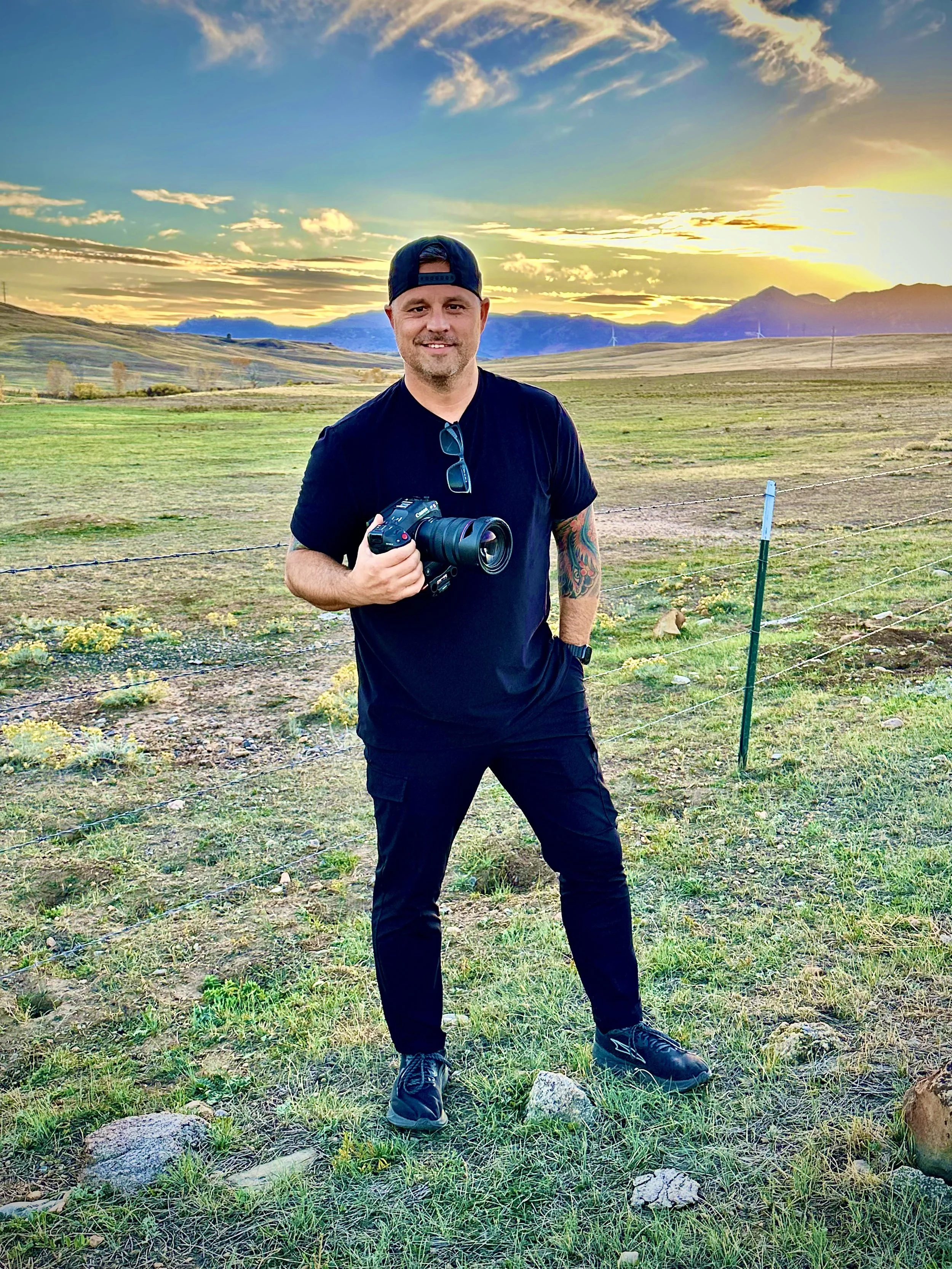 A man standing outdoors in a grassy field holding a camera, wearing a black T-shirt, black pants, and a backward baseball cap, with a sunset sky and mountains in the background.
