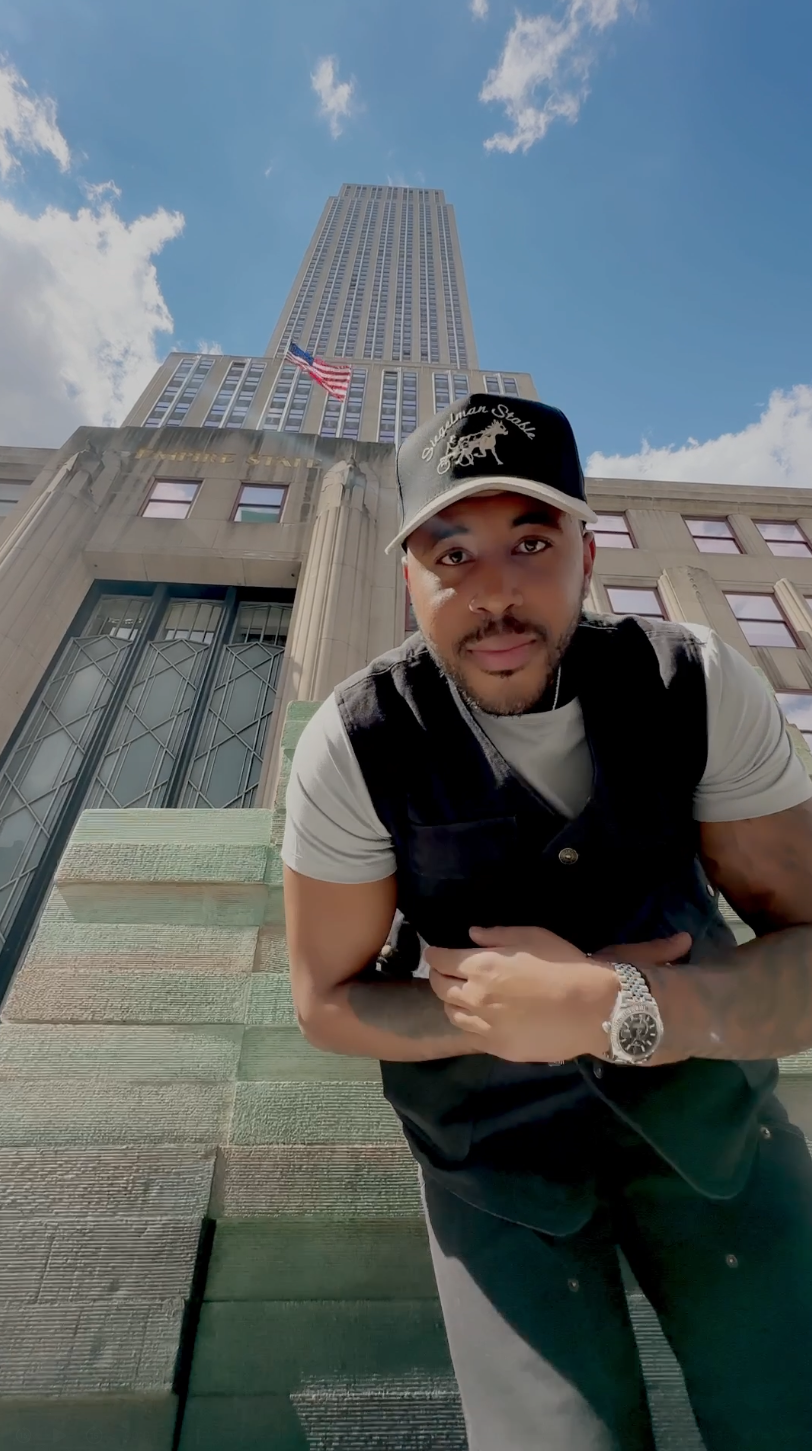 A young man wearing a black and white cap with a horse logo, a white t-shirt, and a black vest, leaning forward with hands crossed in front of him, outdoors in front of a tall skyscraper with an American flag, under a blue sky with some clouds.