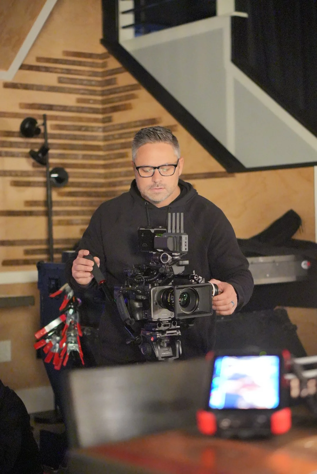 A man wearing glasses and a black hoodie is operating a professional video camera in a studio with wooden wall panels.