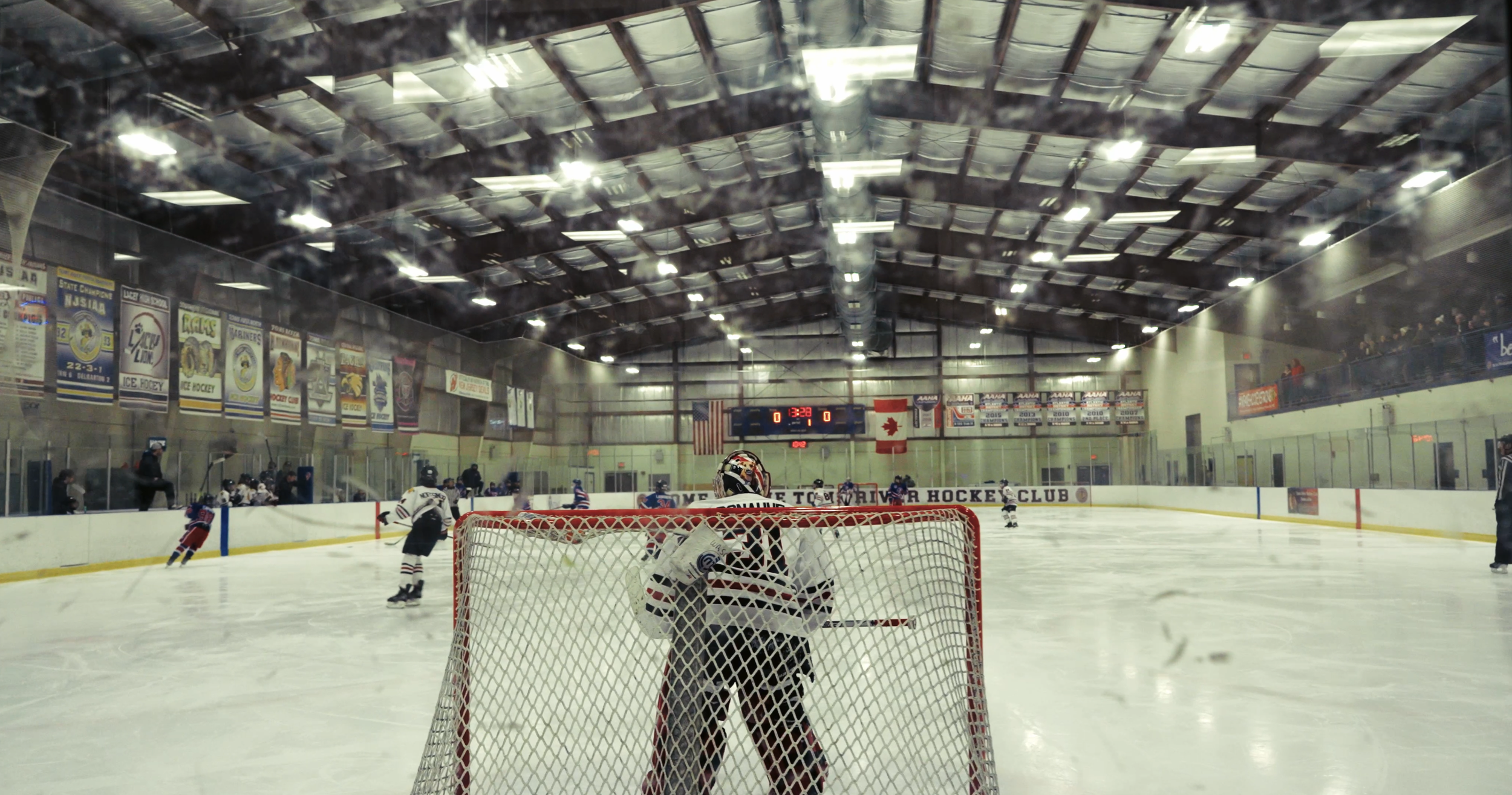 An indoor ice hockey rink with players on the ice, a goalie in front of the net, and spectators on the stands. The ceiling has metallic panels and banners hang along the walls.