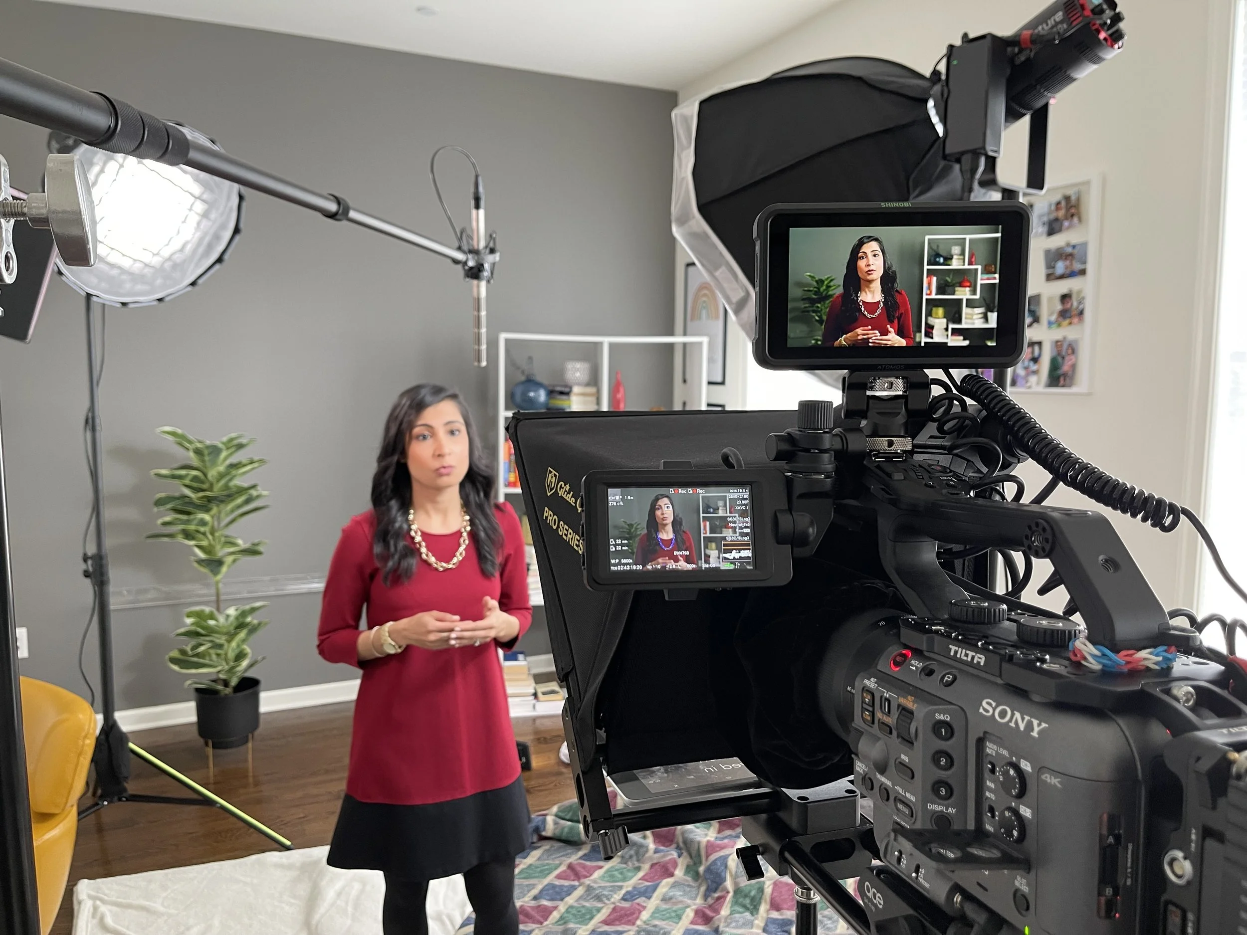 A woman in a red dress is being filmed in a room with a camera setup. Behind her, there is a grey wall, a white shelf with books and decorations, a green plant, and a wall photo collage.