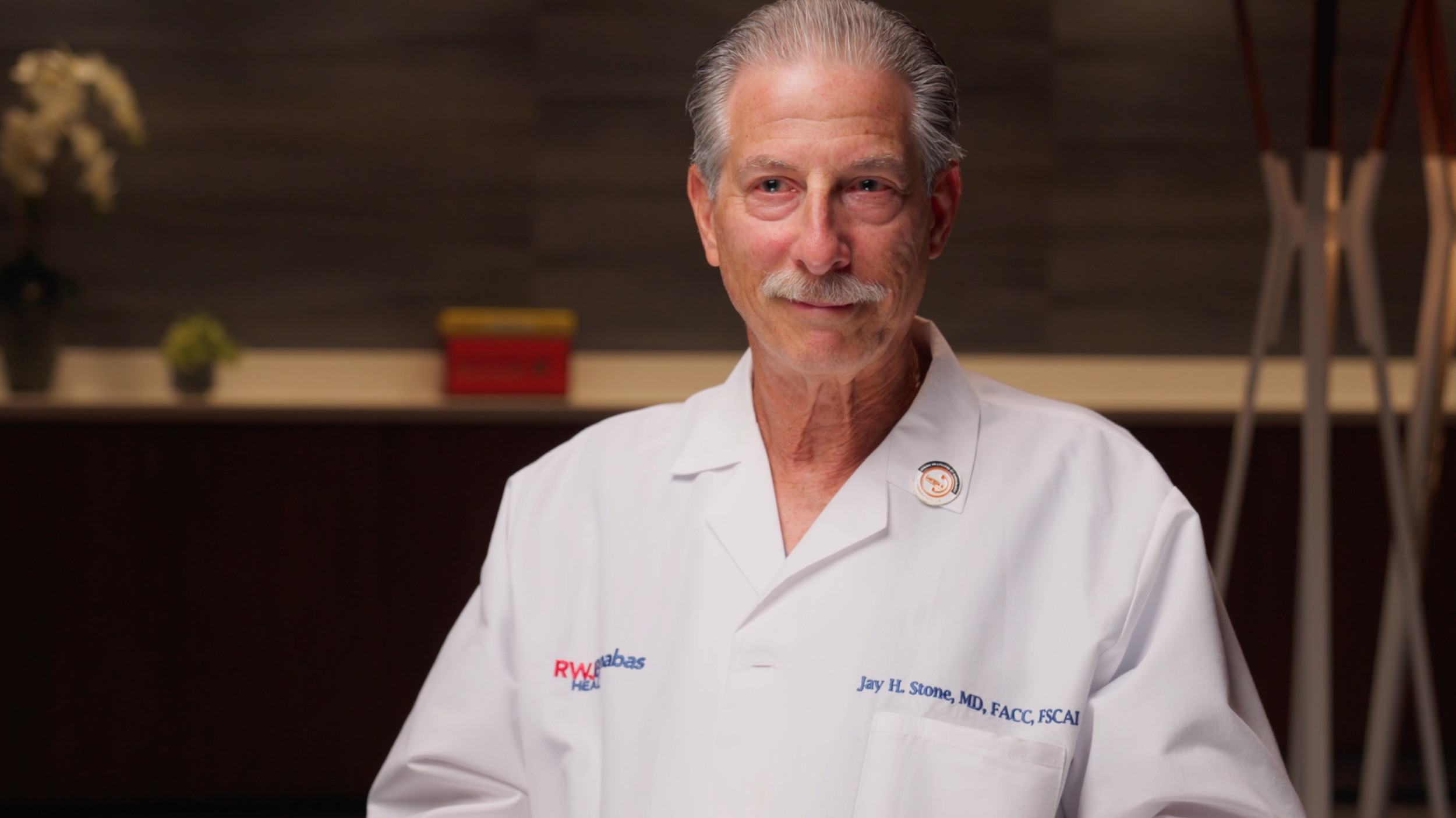 A smiling elderly man with gray hair and a mustache, wearing a white medical coat with a badge that has a red and blue logo. He is standing indoors with blurred background elements including a small potted plant, a red object, and a tall stand.