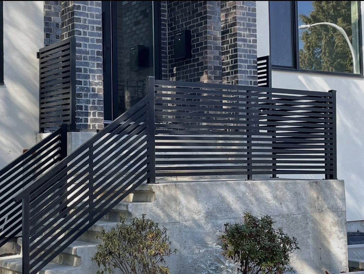 Exterior view of a modern house with a brick wall, glass door, large window, and a black metal railing on a staircase leading to the entrance.