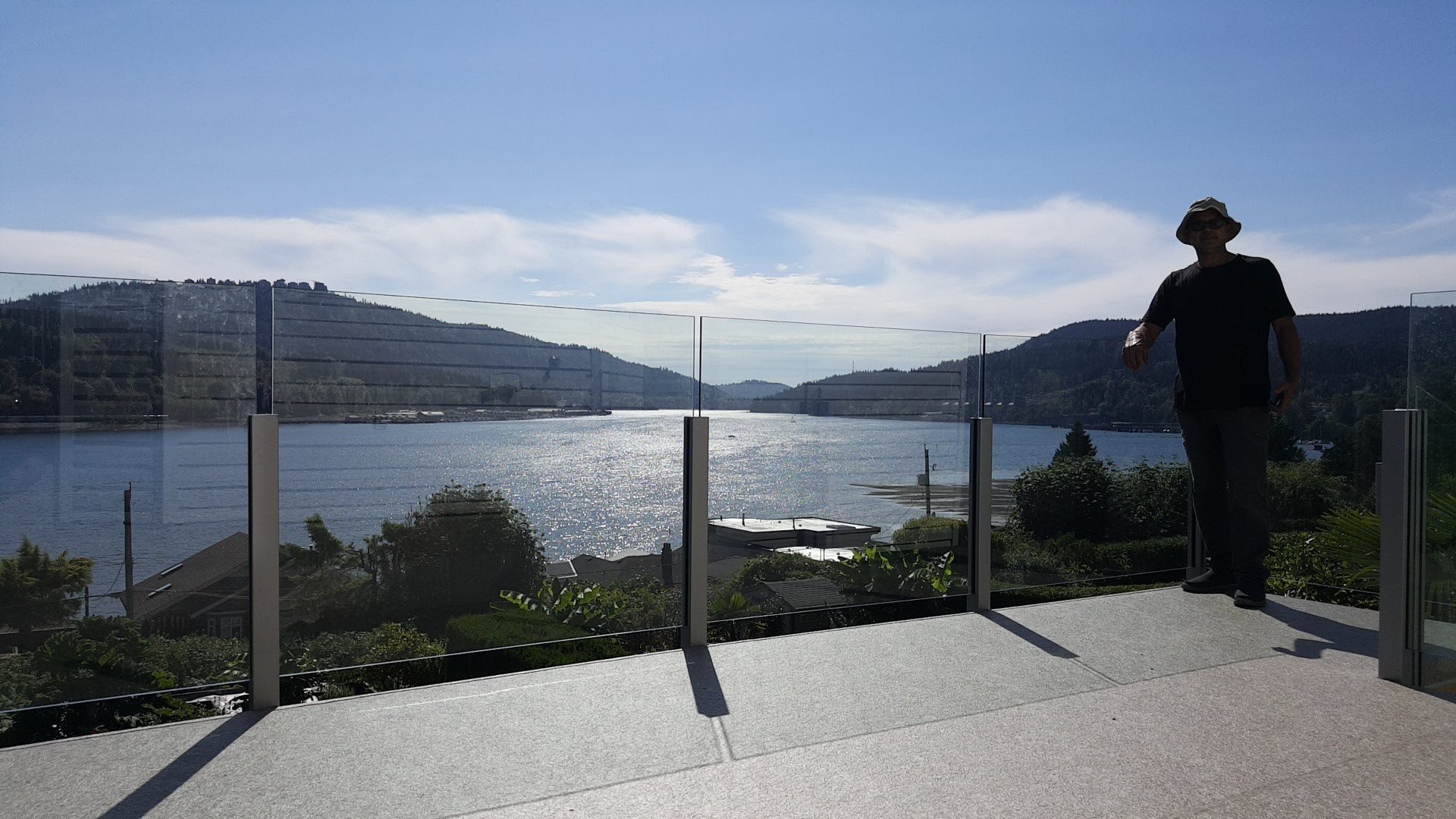 A person standing on a balcony overlooking a large body of water with hills in the background under a partly cloudy sky.