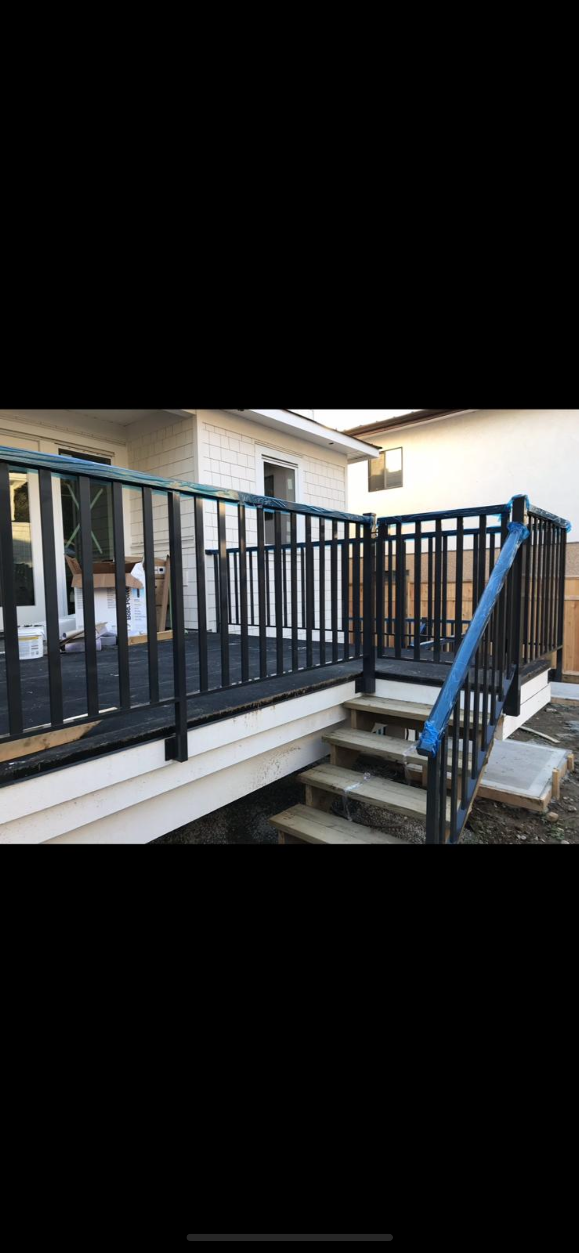 A black metal deck railing installed outside a house, with stairs leading down. The deck is still under construction with some tools and materials visible.