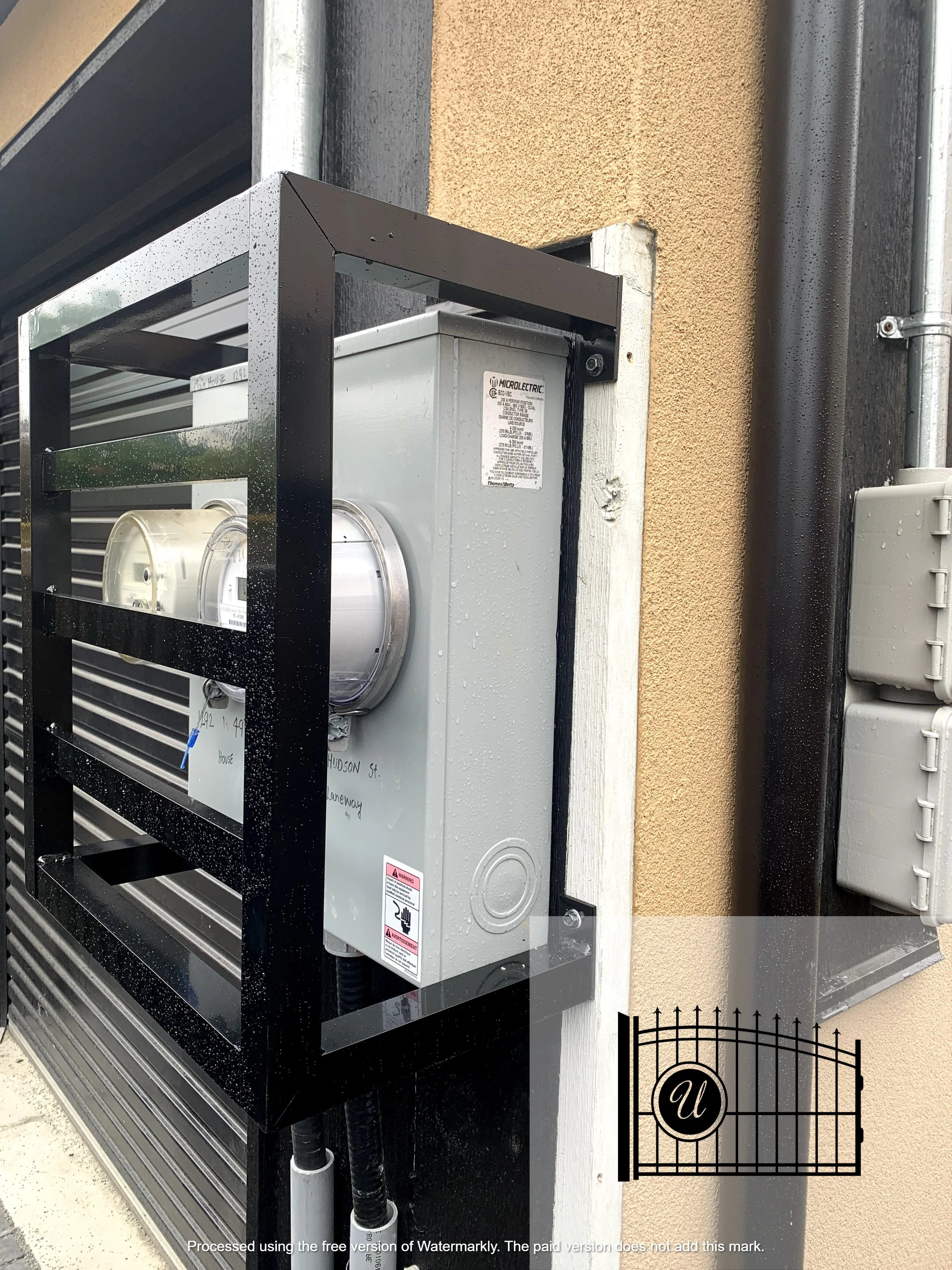 Close-up of an electric utility box mounted on an exterior wall, with black conduit pipes connected underneath, in front of beige stucco wall and surrounding electrical panels.