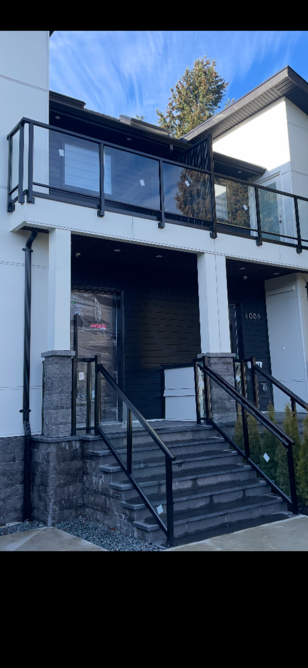 Front view of a modern multi-story house with stairs, glass railing balcony, black and white exterior, and decorative stone elements on the staircase pillars.