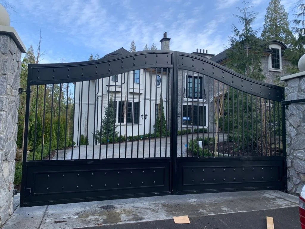 Black metal gate with two sealed sections, in front of a large house with stone and white exterior walls, surrounded by greenery, under a partly cloudy sky.