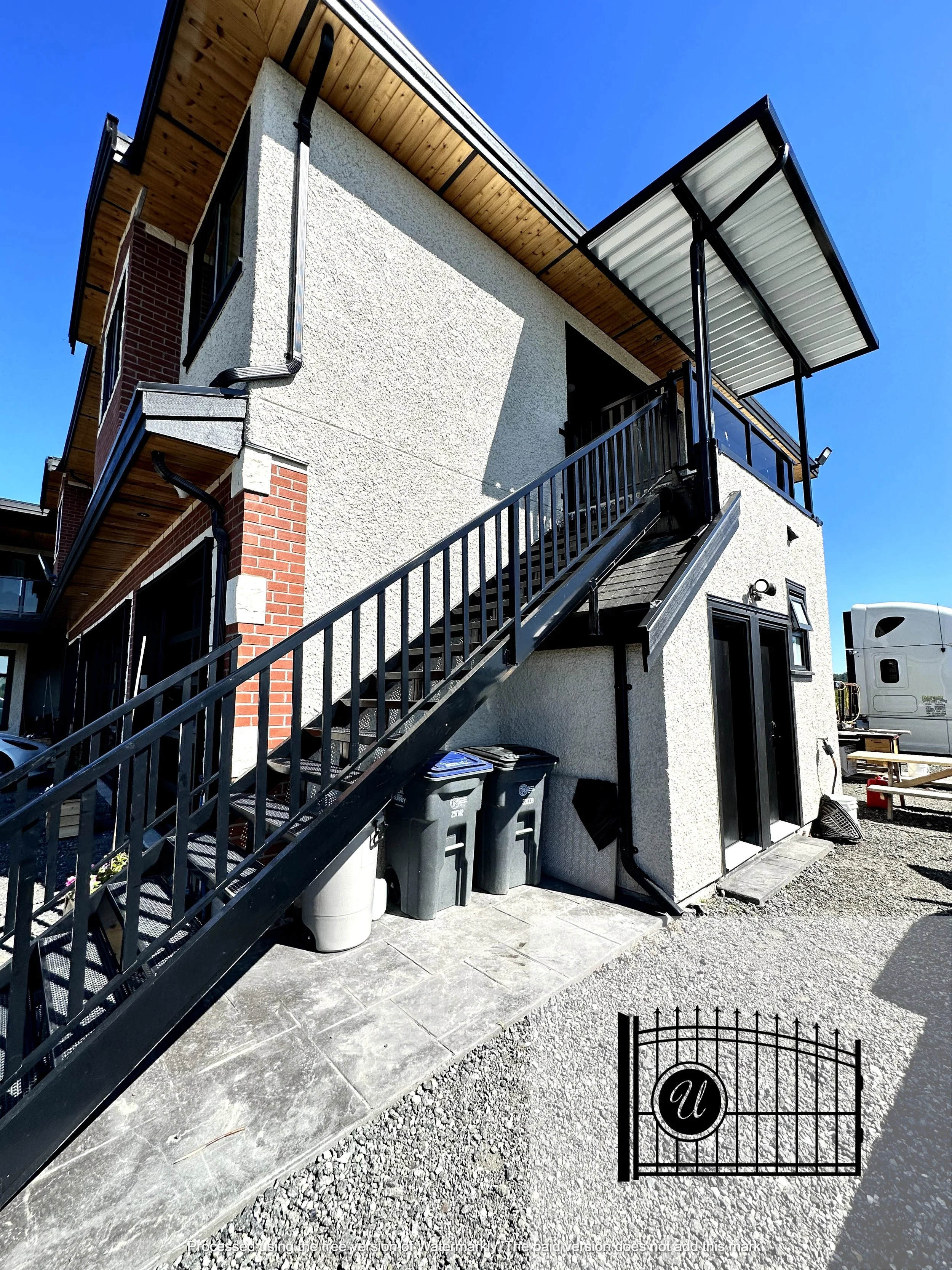 The image shows the exterior of a modern three-story residential building with a stucco and brick façade. It has black metal stairs leading to a balcony with glass railings and a metal roof above. There are multiple large windows, some with black fra