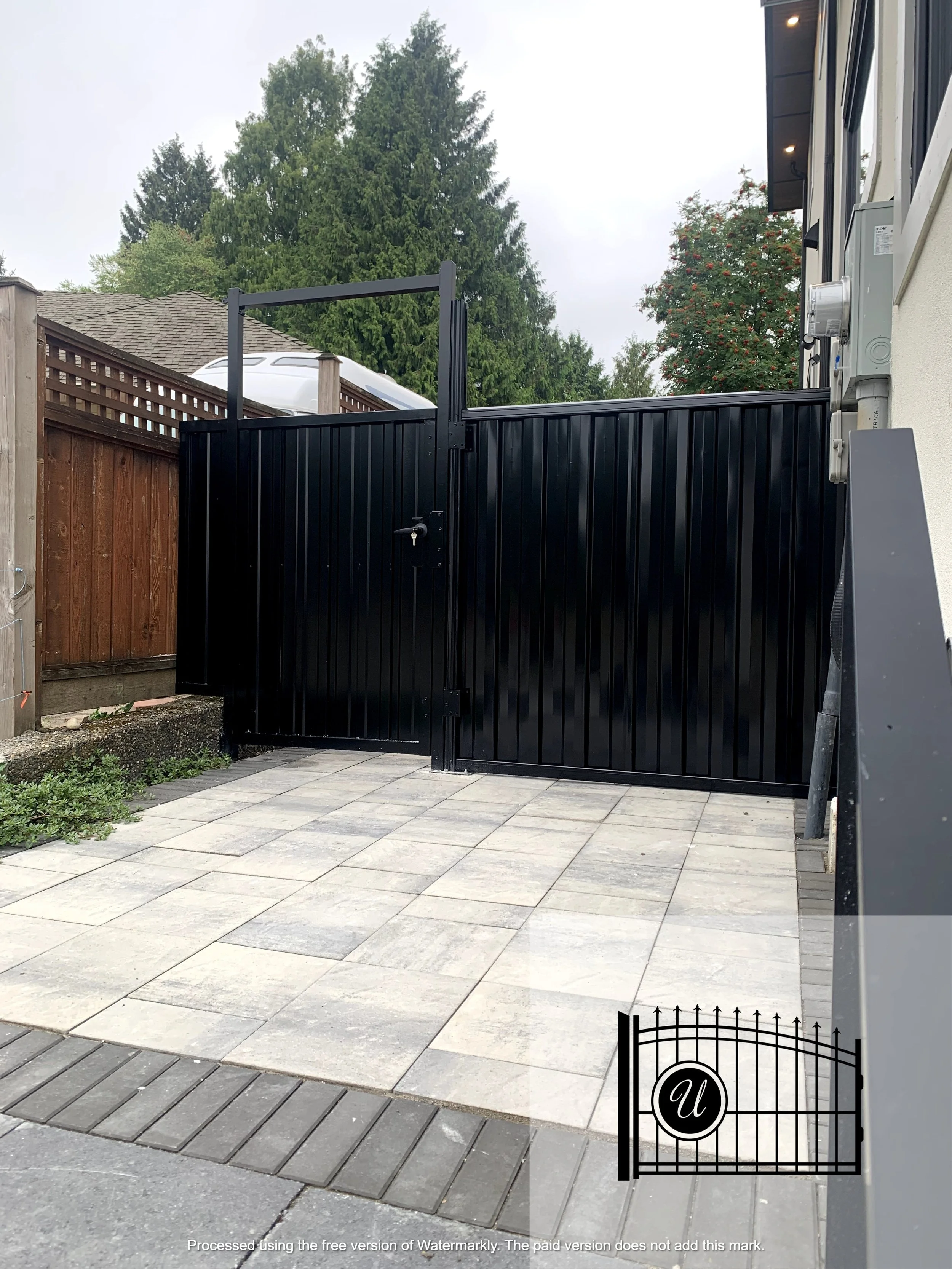 Black metal gate at entrance of backyard with stone tile patio and wooden fencing, house with beige siding and ivy plant visible, trees in background.