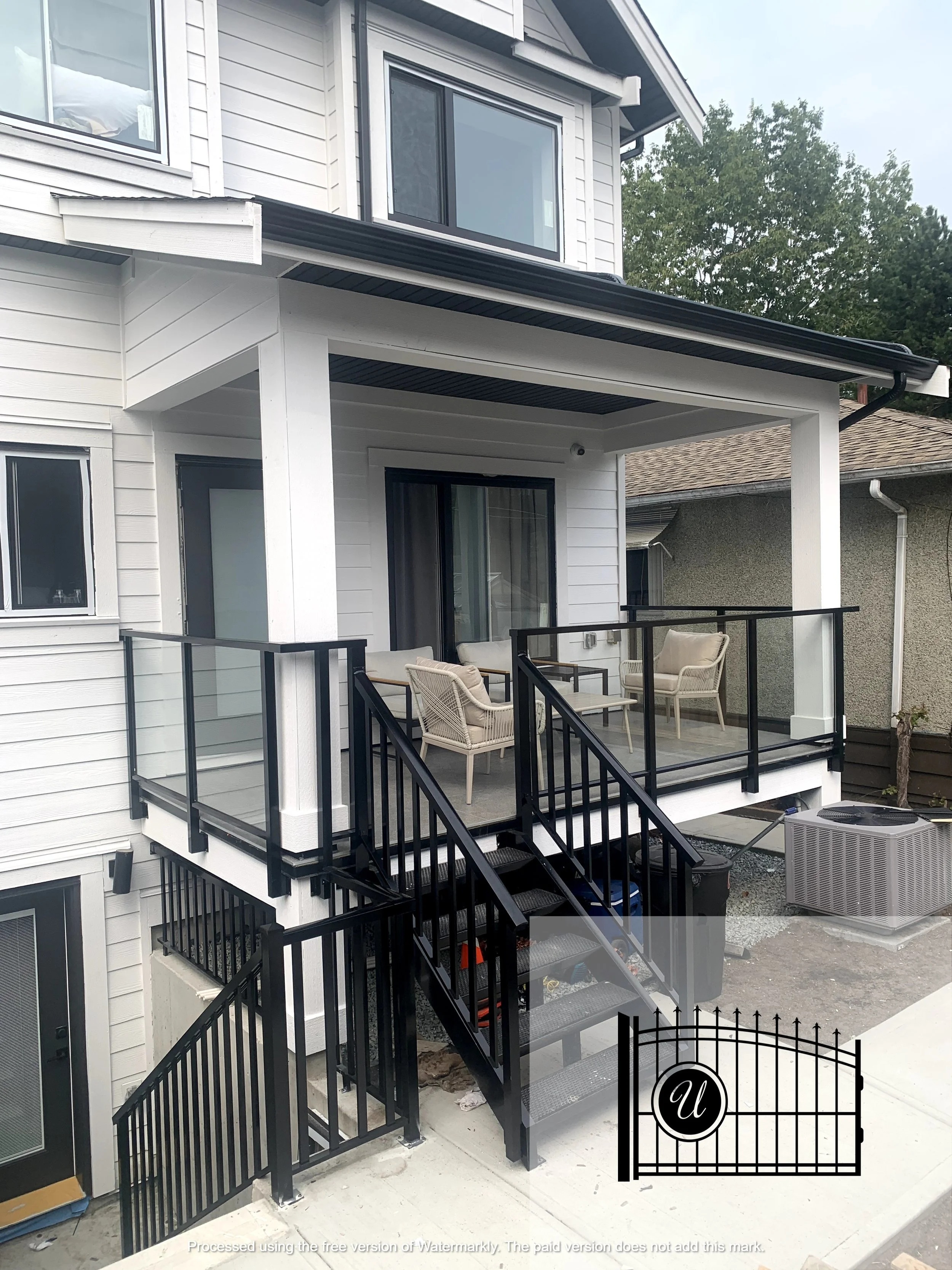 A residential building with a black metal staircase leading to an outdoor balcony with seating and black metal railing. The house has white siding, and there are two sliding glass doors and windows on the upper and lower levels. An air conditioning u