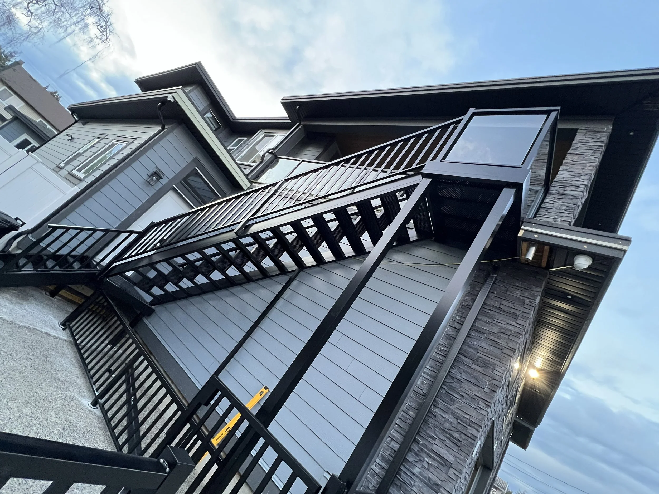 A modern house with gray siding, black accents, and a stone wall, featuring a metal fire escape staircase on the exterior, against a background of a partly cloudy sky.