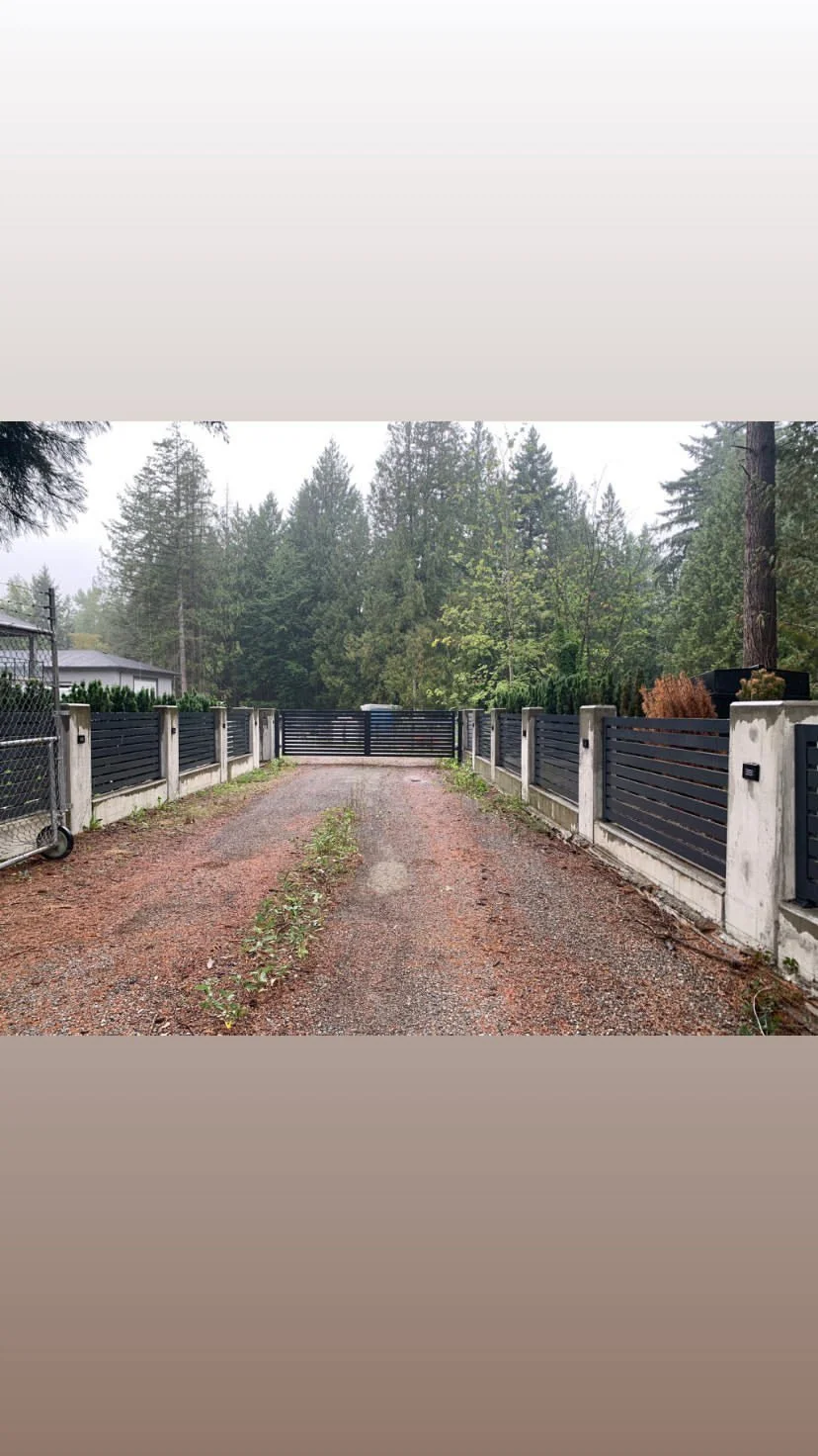A gravel driveway with weeds running through the middle, flanked by a modern black fence and concrete posts, leading to a closed black gate surrounded by trees in a wooded area.