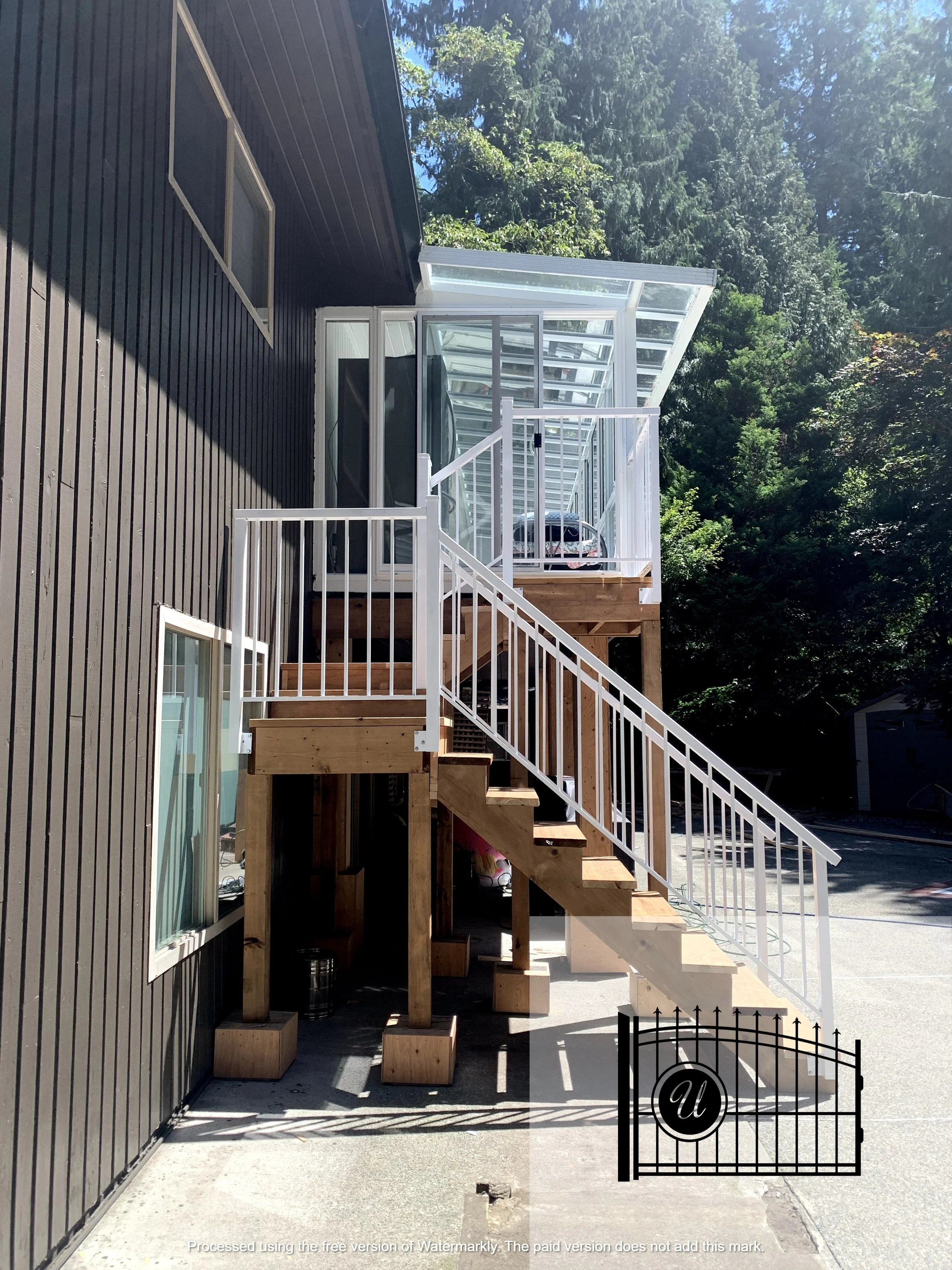 Backyard with a black exterior house wall, a staircase with white railings leading to a small deck with a glass enclosed sunroom, surrounded by tall trees, and a small black iron gate.