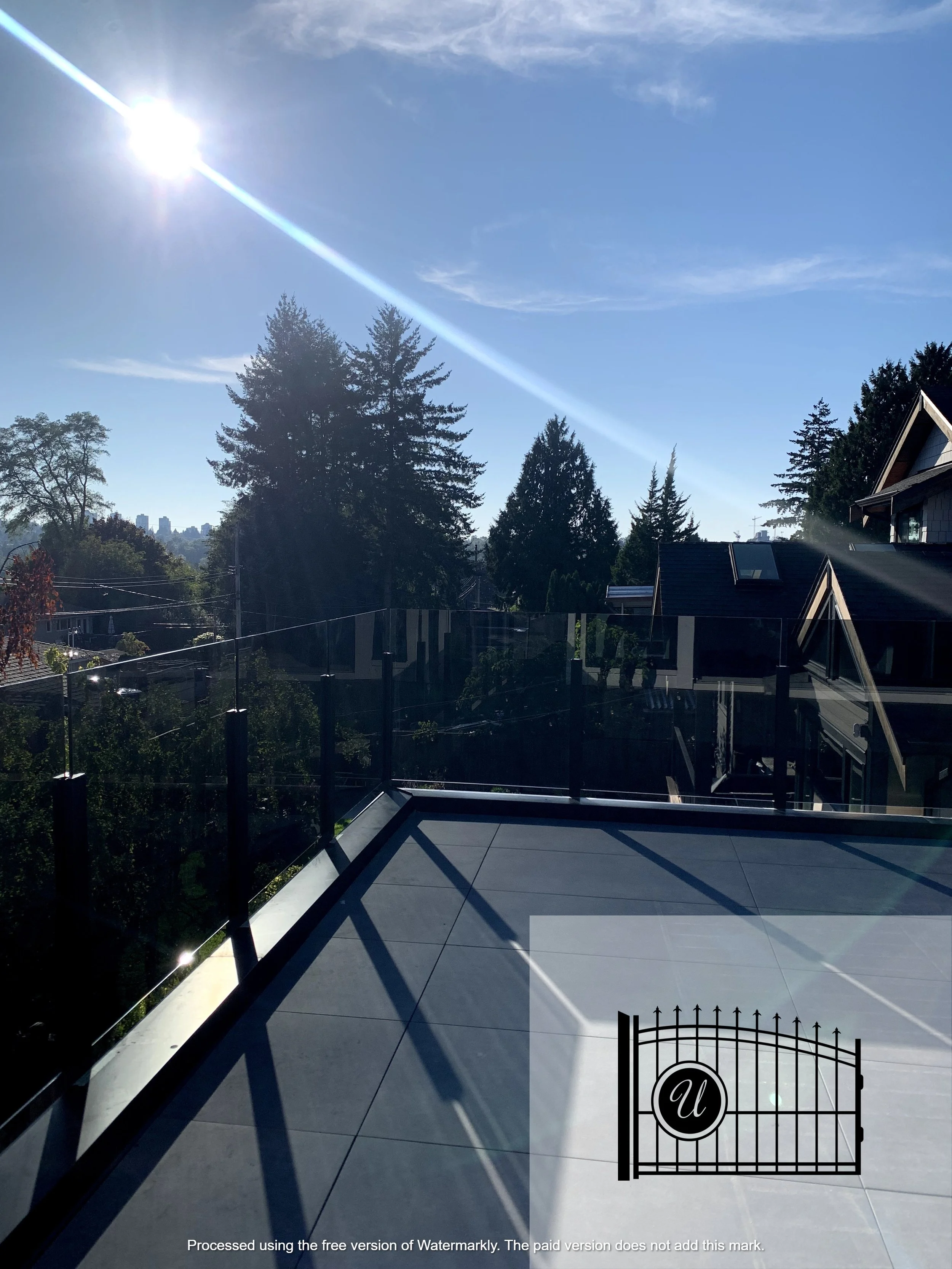 A view from a balcony showing a black iron gate with a round emblem that has the letter 'U,' overlooking trees, rooftops, and a city skyline in the distance under a clear blue sky with some clouds and the sun shining brightly.